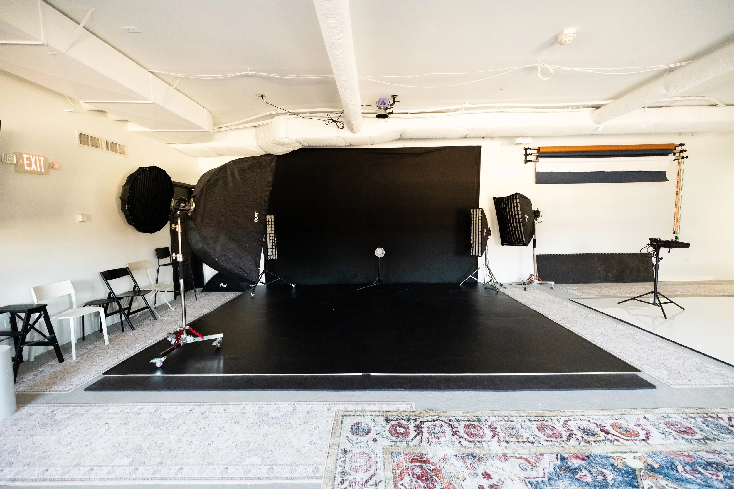 Photography studio setup with black backdrop, studio lights, and a tripod on a black floor, surrounded by white walls, chairs, and a patterned rug.