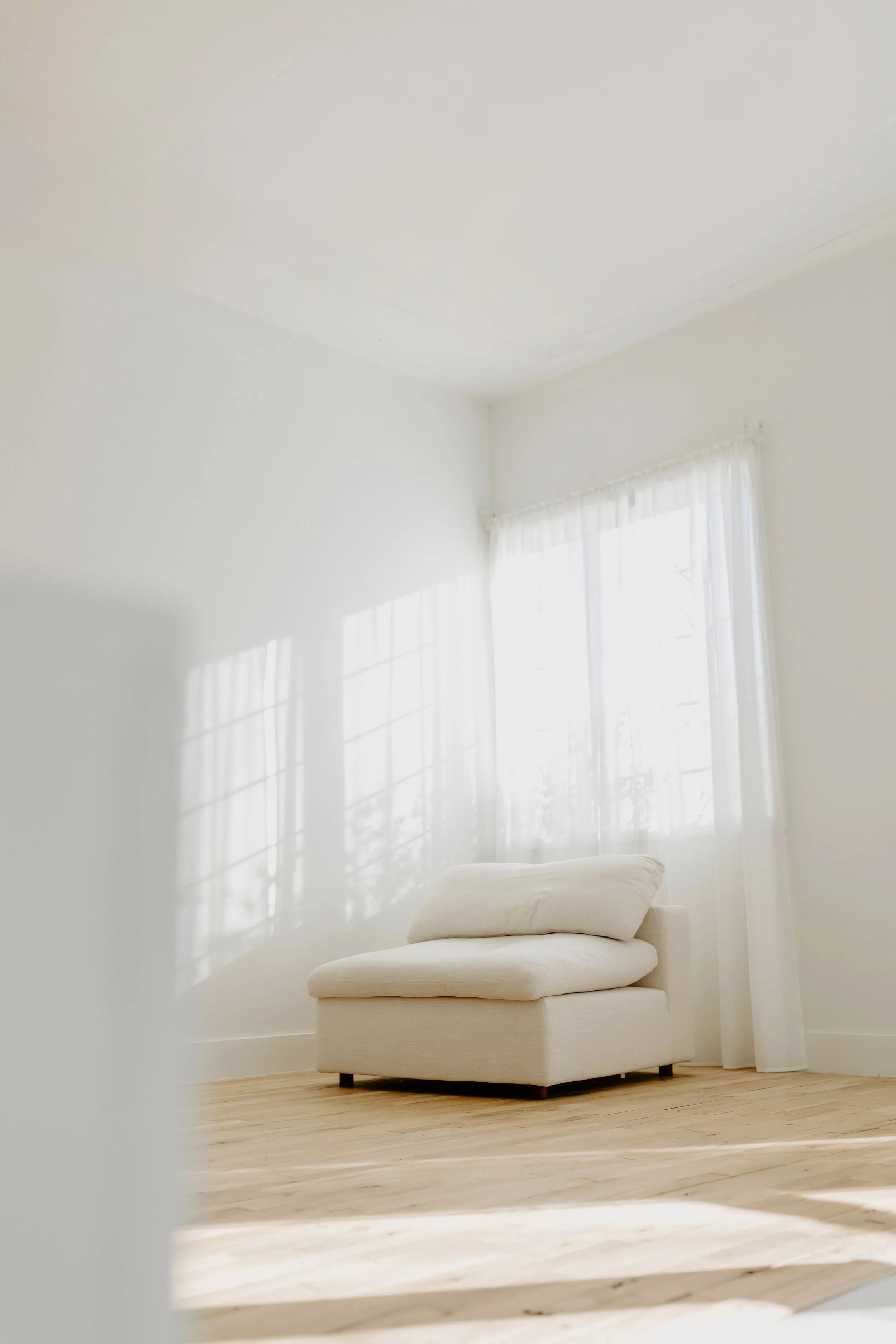 A minimalistic, bright living room corner with a white armchair near a window with sheer white curtains, soft sunlight casting shadows on the wall and floor.
