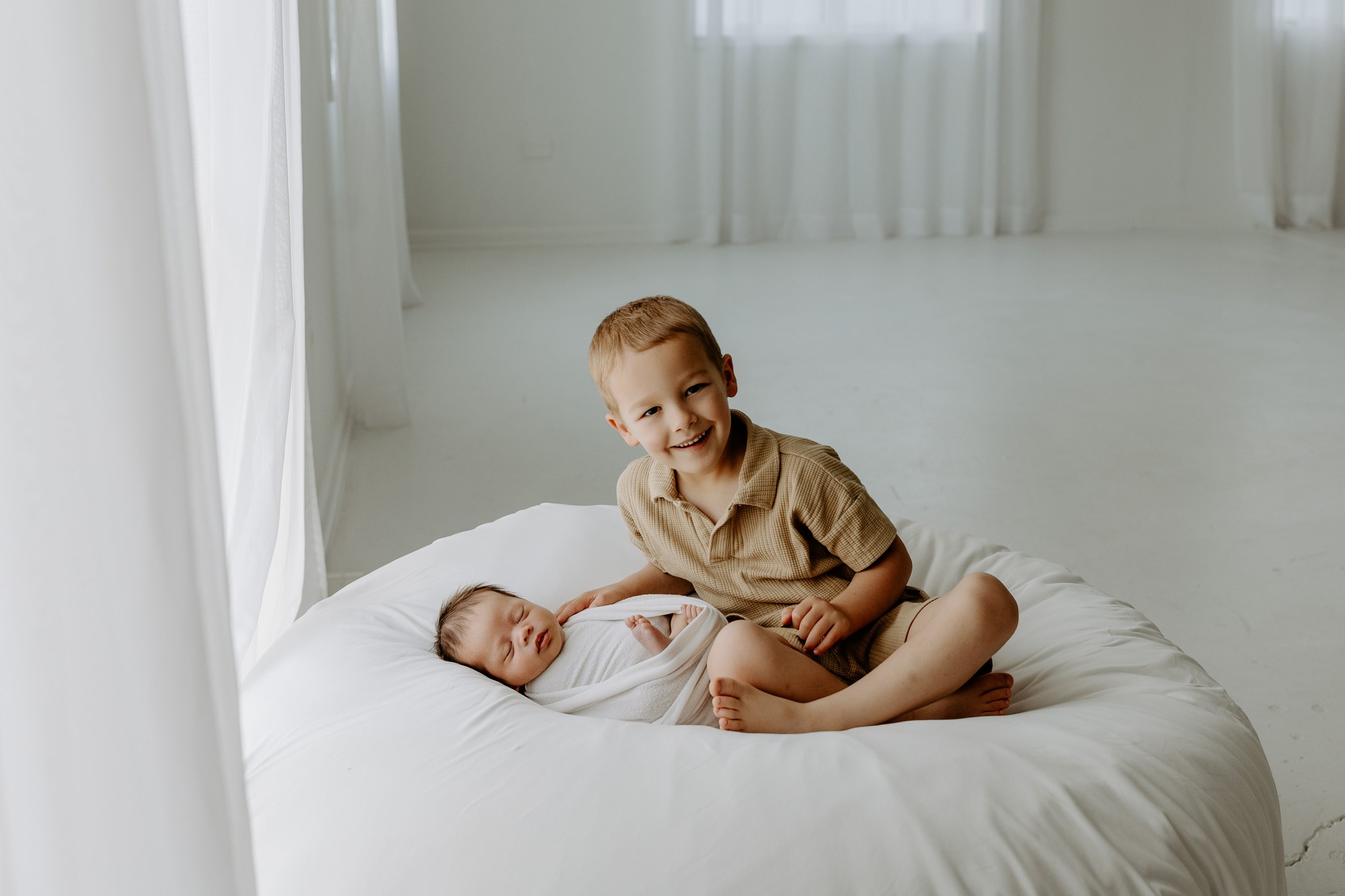 A young boy sitting on a bed next to a sleeping baby, both smiling, in a bright, airy room with white curtains.