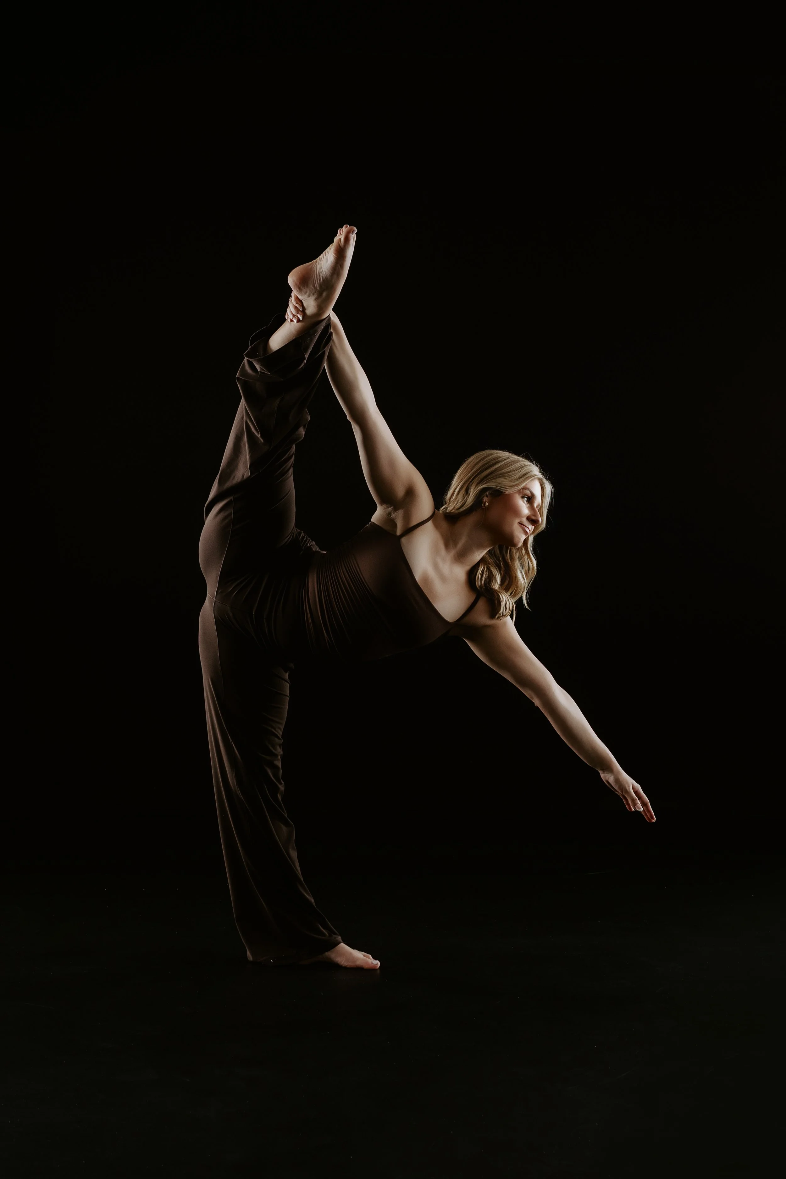 A woman with long blonde hair practicing yoga against a black background, wearing dark athletic clothing, performing a high leg stretch with one arm extended downward.
