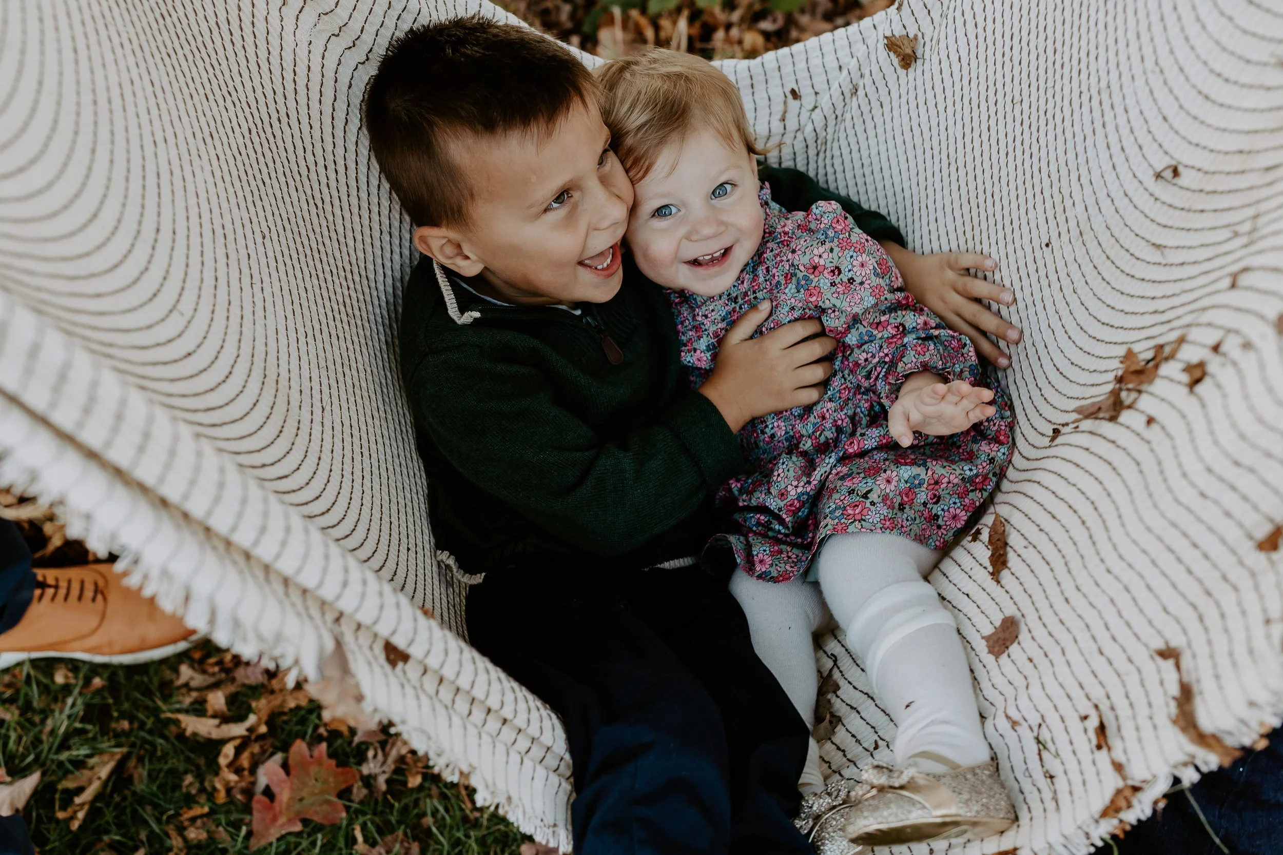 Two young children, a boy and a girl, sitting on a striped outdoor couch with fallen leaves around them. The boy has short dark hair and is wearing a dark sweater. The girl has light hair and is wearing a floral dress with white tights. They are smil