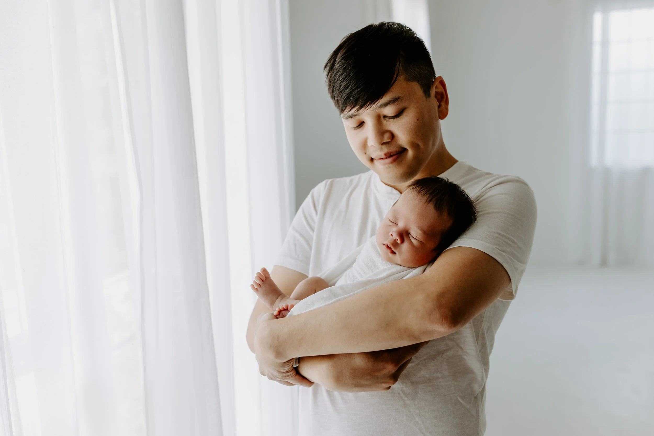A man holding a sleeping baby near a window with white curtains.