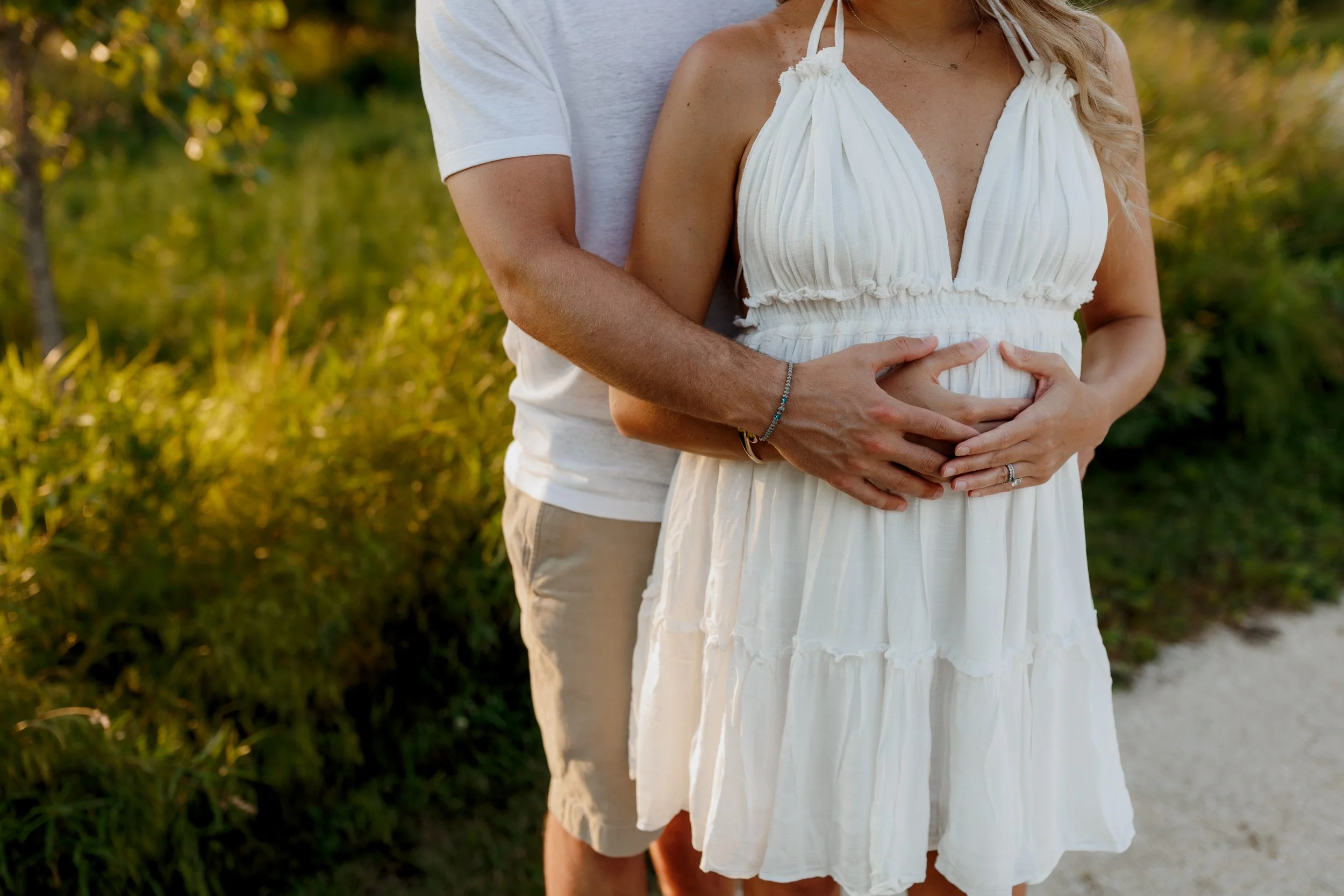 A pregnant woman in a white dress being embraced from behind by a man, with greenery in the background, during sunset.
