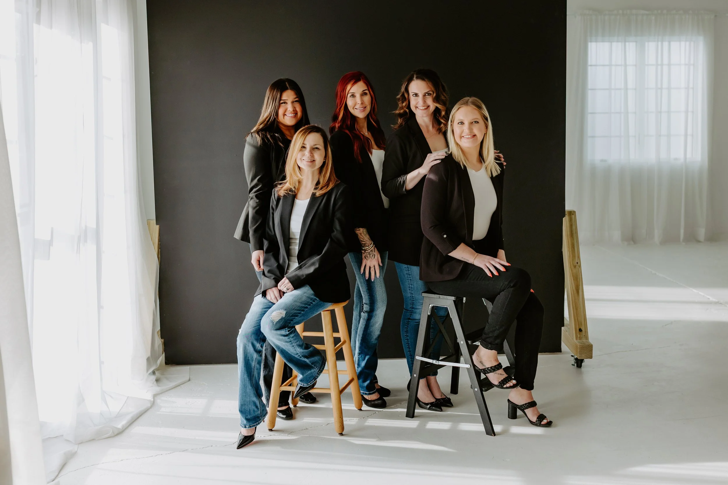 Group of six women dressed in black blazers and jeans, posing together in a photo studio with a black background and white curtains.
