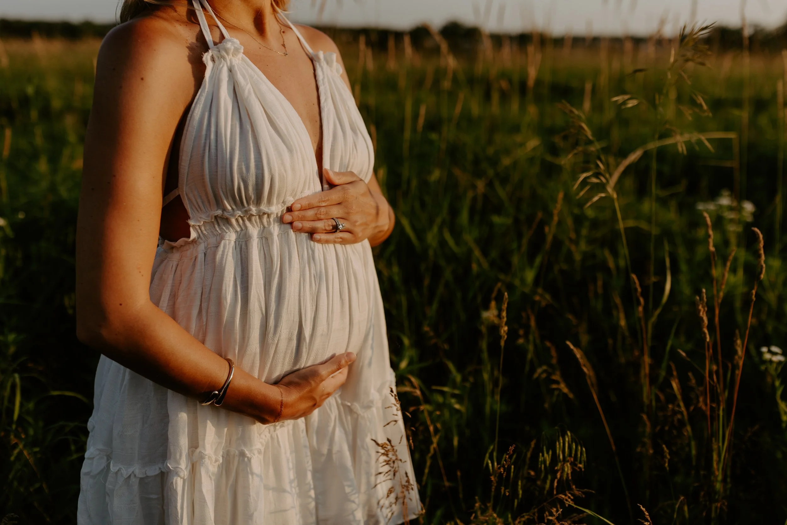 A pregnant woman in a white dress stands in a field of tall grass during sunset, gently cradling her belly with both hands.