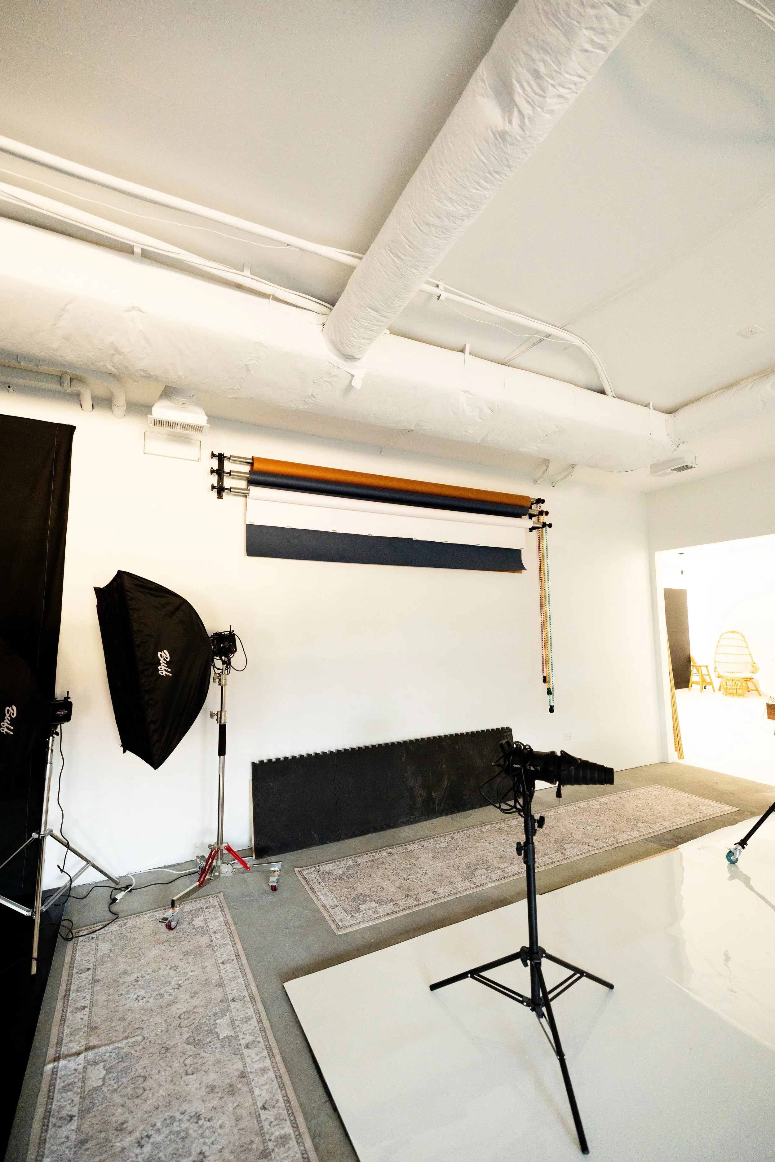 Photograph of a photography studio with lighting equipment, backdrop rolls, and a camera on a tripod, set up on a white floor with rugs, in a brightly lit room with white walls and ceiling.