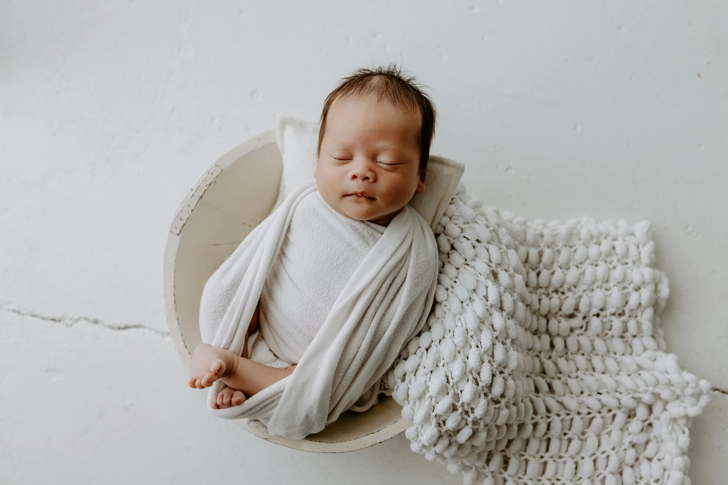 A sleeping baby wrapped in white cloth, resting on a soft, textured white blanket, inside a round, white container against a plain white background.