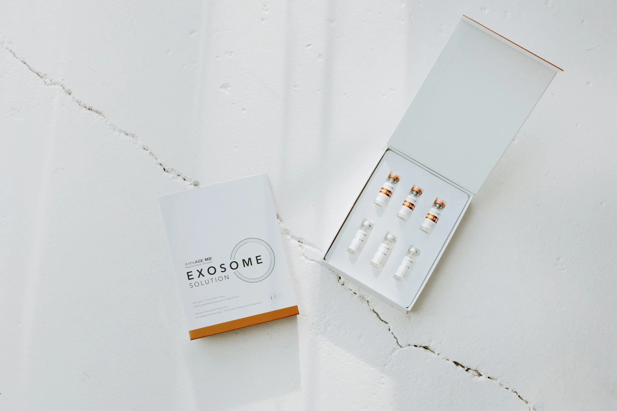 A white box labeled EXOSOME Solution containing small vials, with an open lid revealing six vials inside, and a brochure beside it, all placed on a white surface.