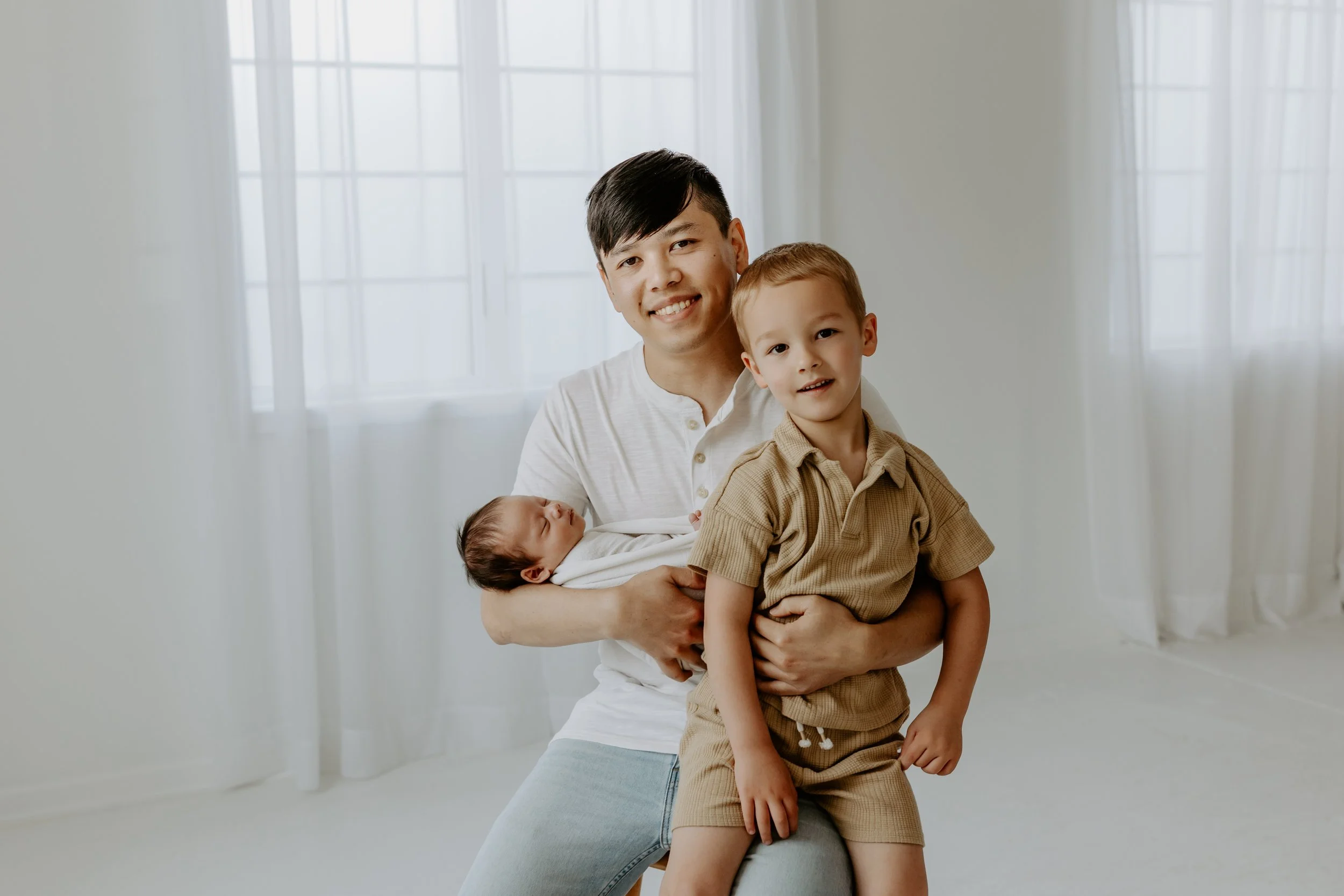 A man with black hair holding a young boy with blonde hair, who is sitting on his lap, while a newborn baby sleeps in his arm. The background shows large white curtains and windows with natural light.