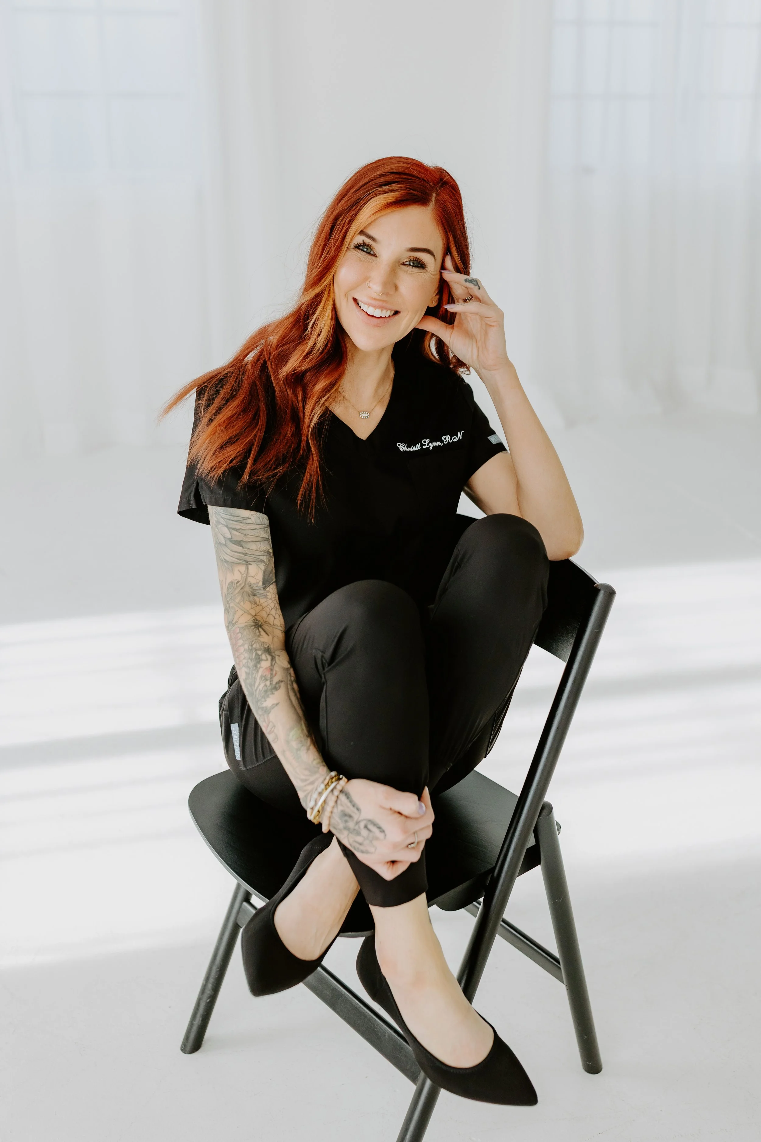 A cheerful woman with red hair and tattoos on her left arm, sitting on a black chair in a bright, minimalistic room, smiling at the camera.