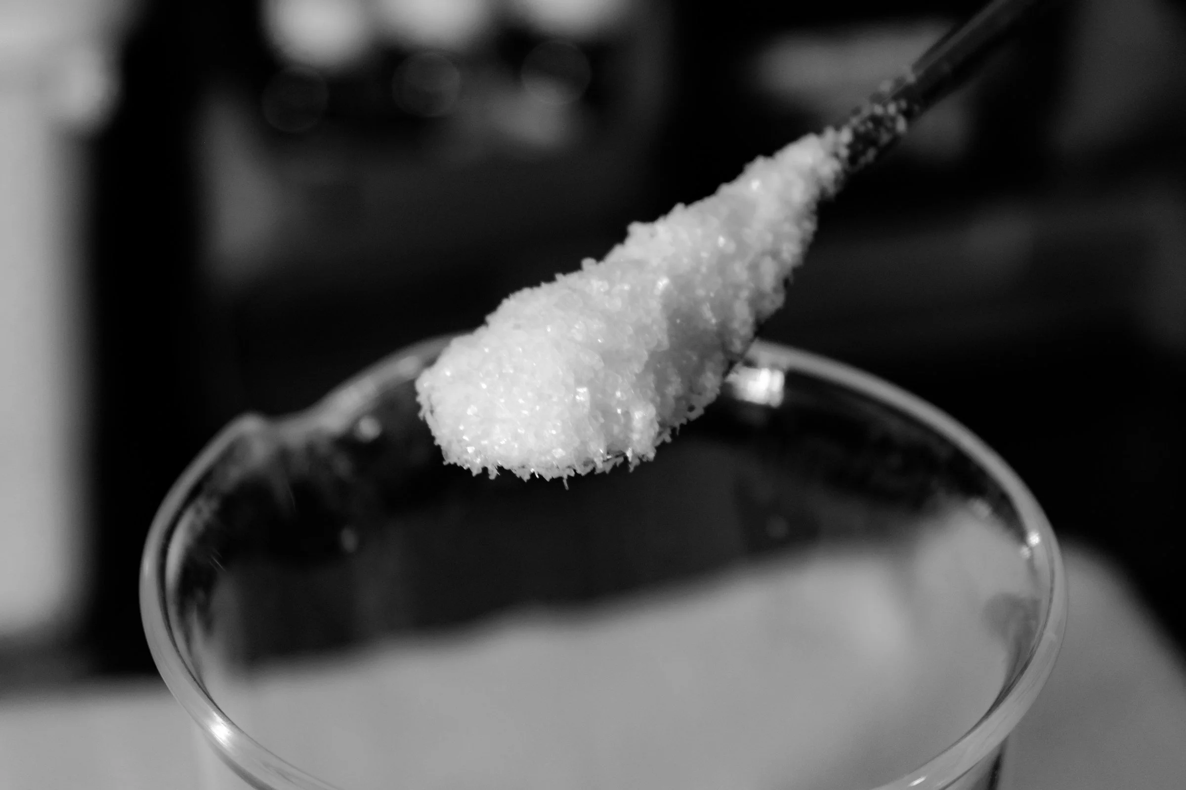 A bowl with a spoonful of sugar being added over a glass of liquid, in black and white.