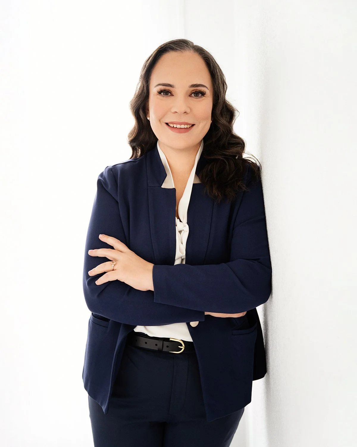A woman in a navy suit with a white blouse, smiling, standing with arms crossed against a plain light background.