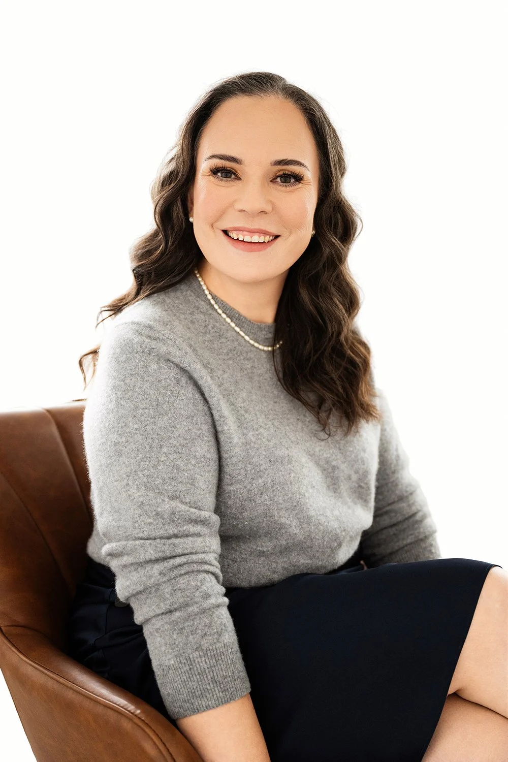 A woman with long wavy brown hair, wearing a gray sweater, pearl necklace, and black skirt, sitting and smiling against a white background.