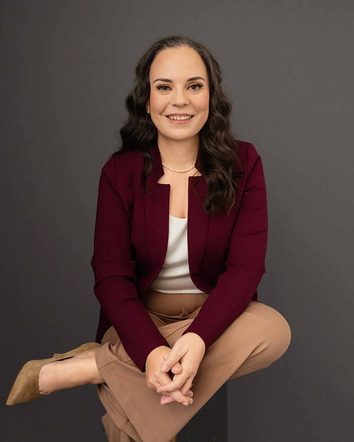 A woman with long, wavy dark hair and light skin, smiling and sitting against a dark grey background. She is wearing a maroon blazer, a white top, tan pants, and tan shoes. She is accessorized with pearl earrings and a pearl necklace.