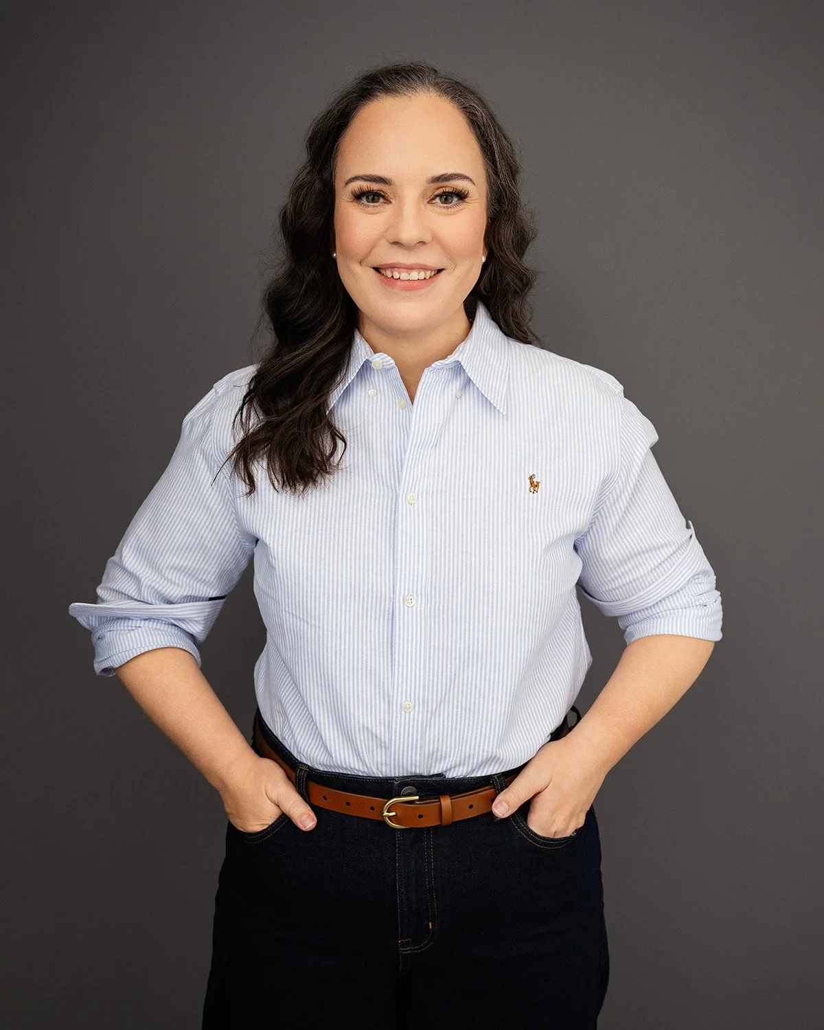 Portrait of a woman with dark hair wearing a white striped button-up shirt with rolled sleeves, black pants, and a brown belt, standing against a dark gray background.