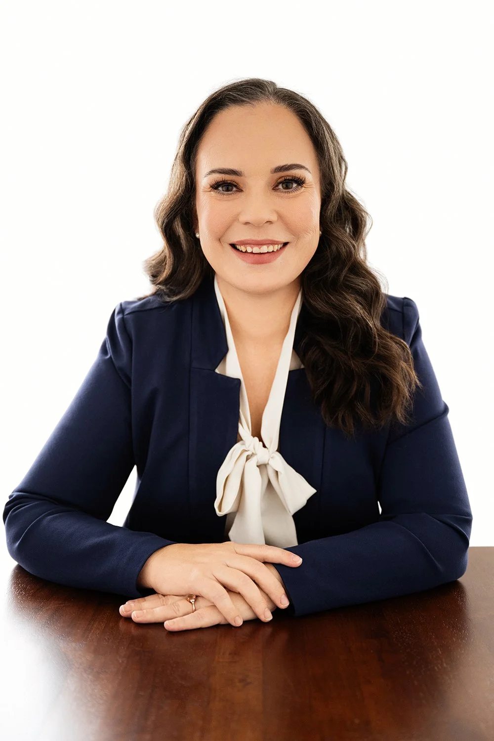 A smiling woman with wavy brown hair sitting at a dark wooden table, wearing a navy blazer and a white blouse with a bow tie.