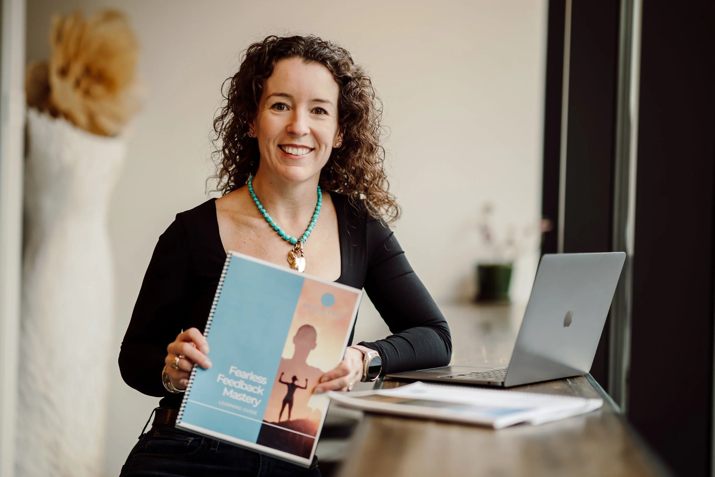 Image of a woman sitting near her laptop holding a booklet with the words Fearless Feedback Mastery on the cover