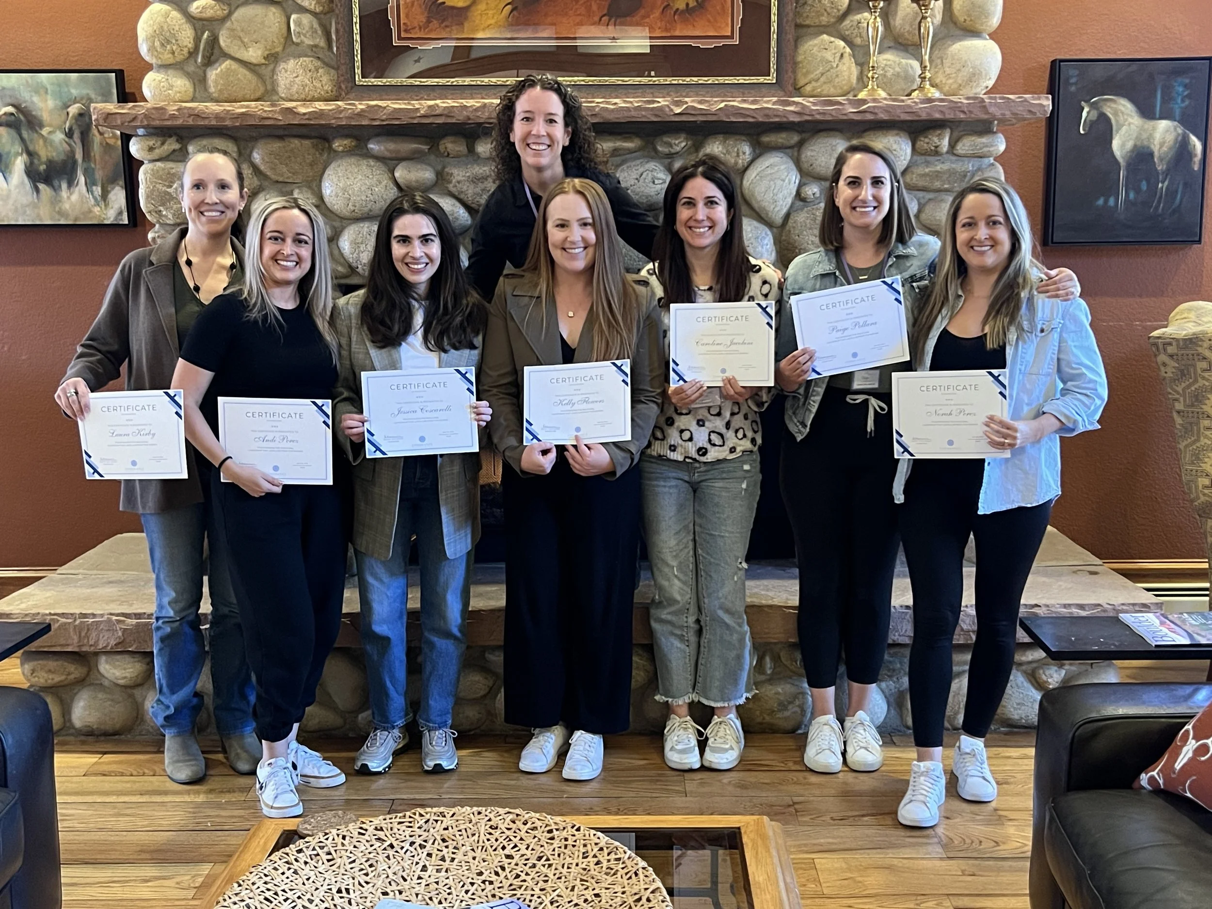 An image of 8 women in front of a river rock fireplace, each holding a certificate of completion for the Leadership That Lasts: A Retreat For Women.