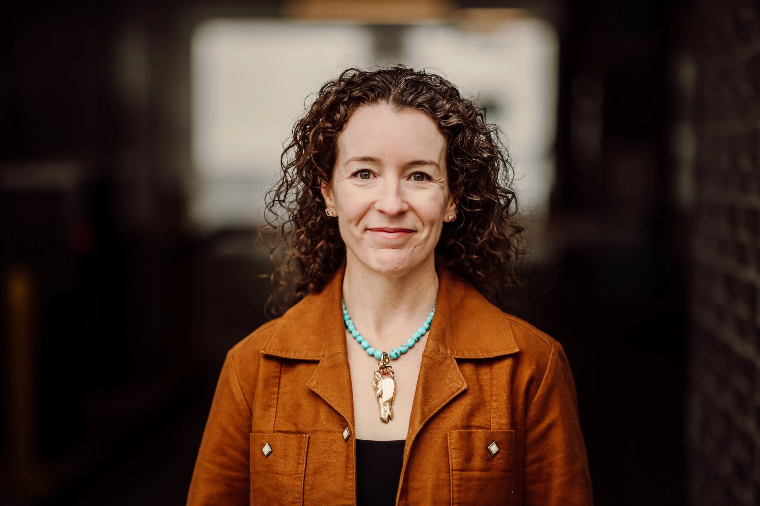 A woman with curly brown hair, wearing a beige scarf and a denim jacket, standing outdoors near a dark background, smiling softly at the camera. Katie is an Executive Coach, founder of Endurance Management Coaching.