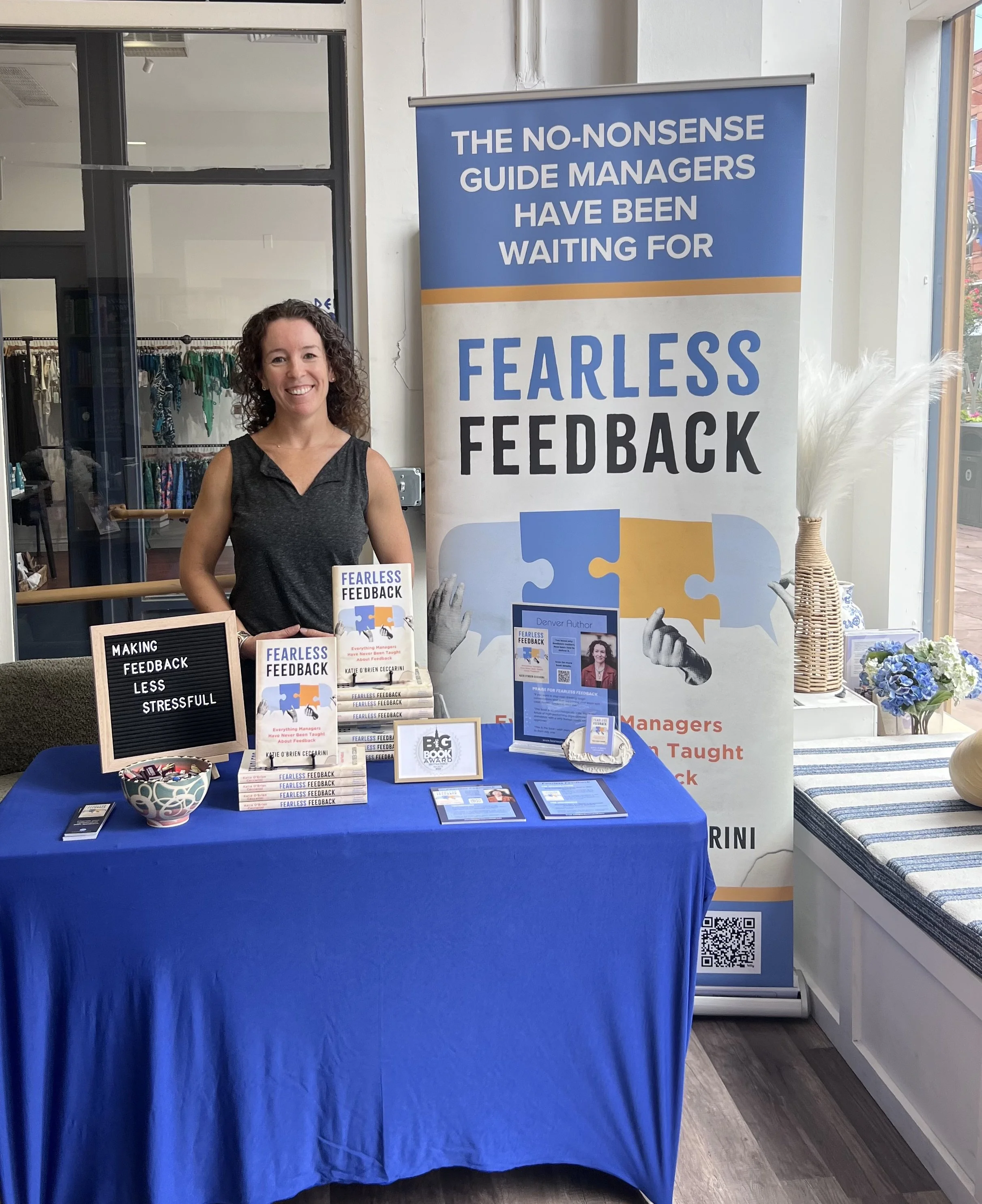 This is a photo of Katie O'Brien Ceccarini at an author event, standing in front of an expandable banner with the cover of Fearless Feedback on it. Fearless Feedback recently won 2026 NYC Big Book Distinguished Favorite in leadership.
