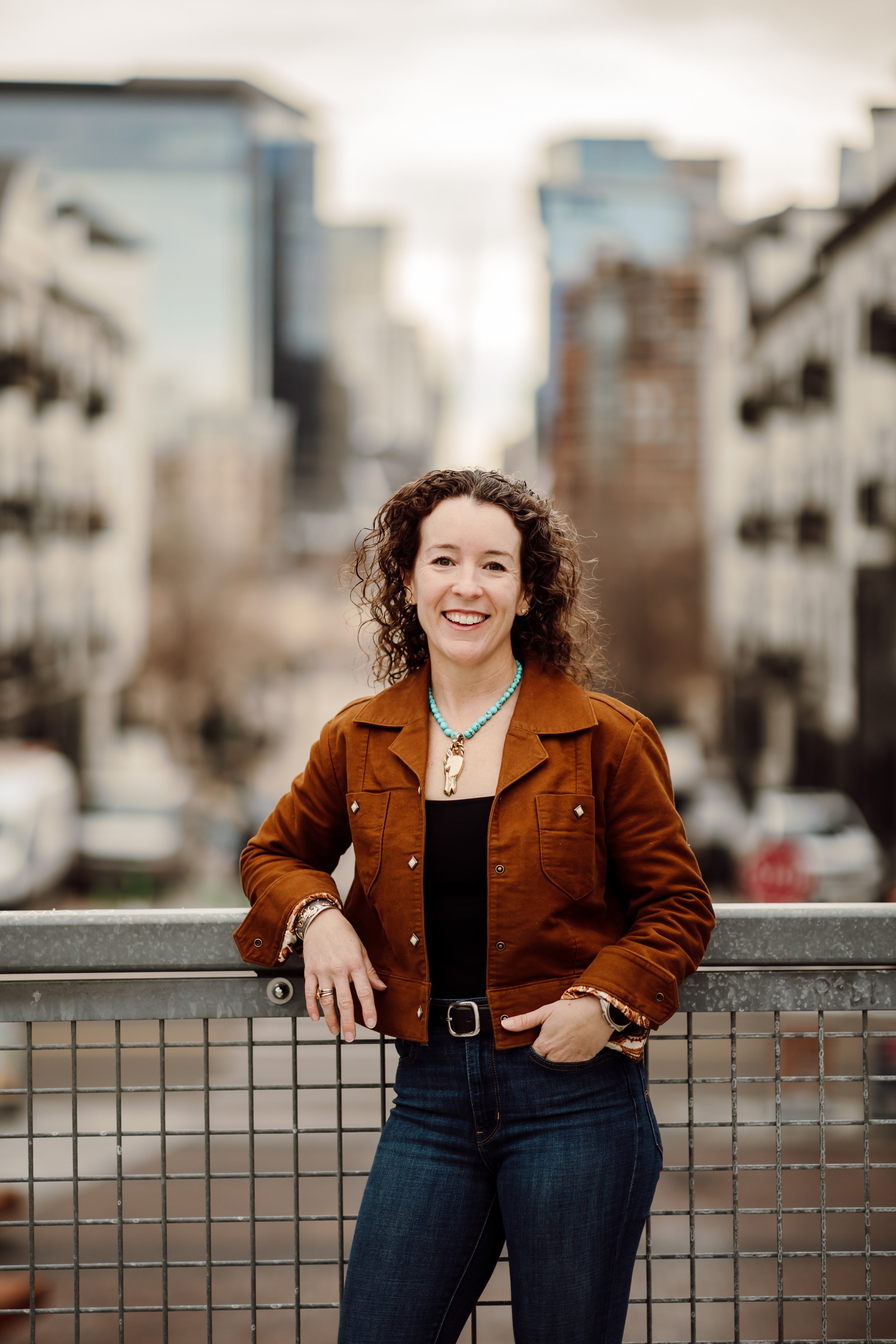 Image of a woman standing against a fence with city buildings behind. She's wearing dark pants, a rust colored jacket, and has a big smile.