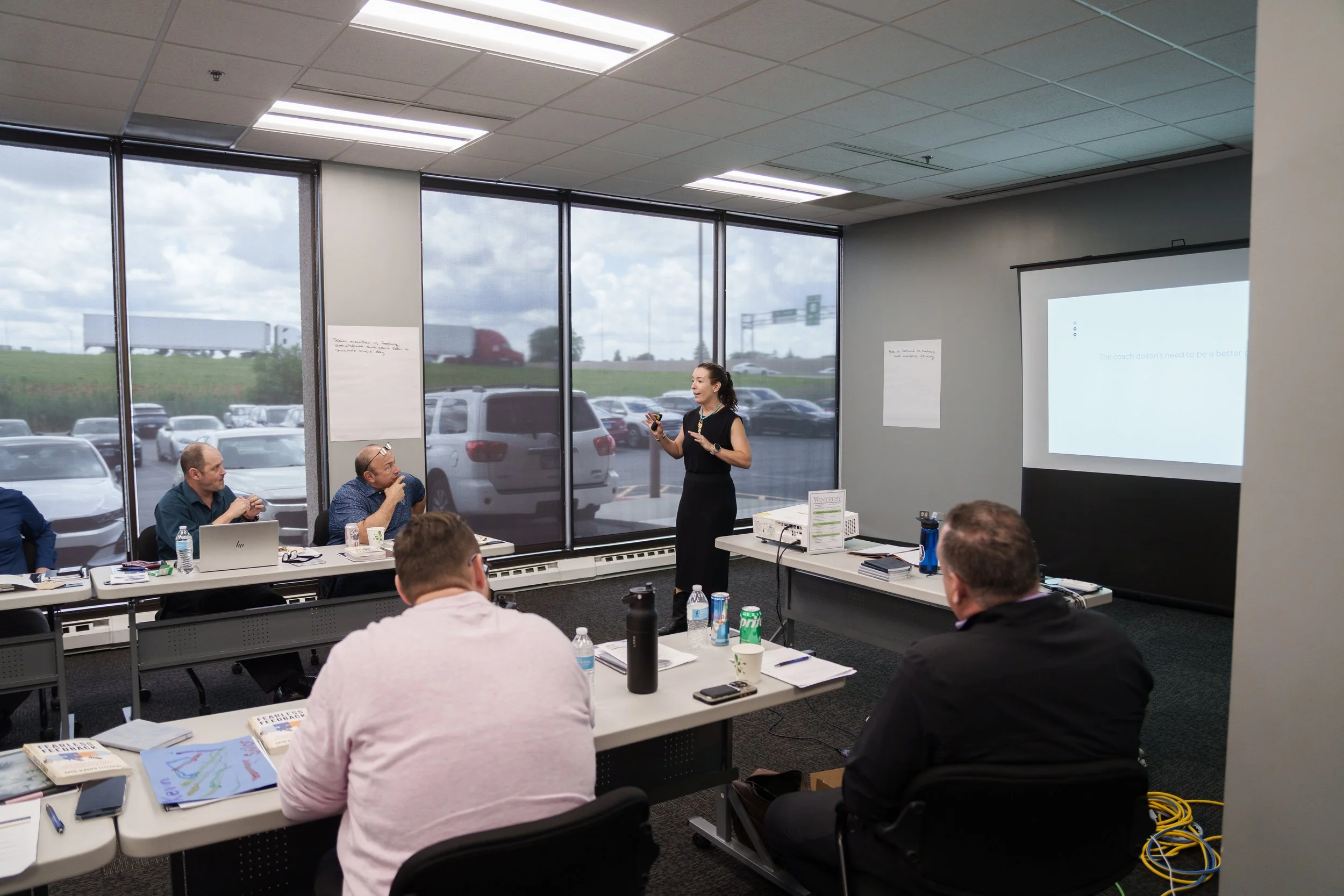 An image of Katie O'Brien Ceccarini at the front of a room of AllState Insurance agency owners and managers, teaching them a 3 hour workshop on Managers as Coaches. There's a large screen with content projected and you can see tables of people.