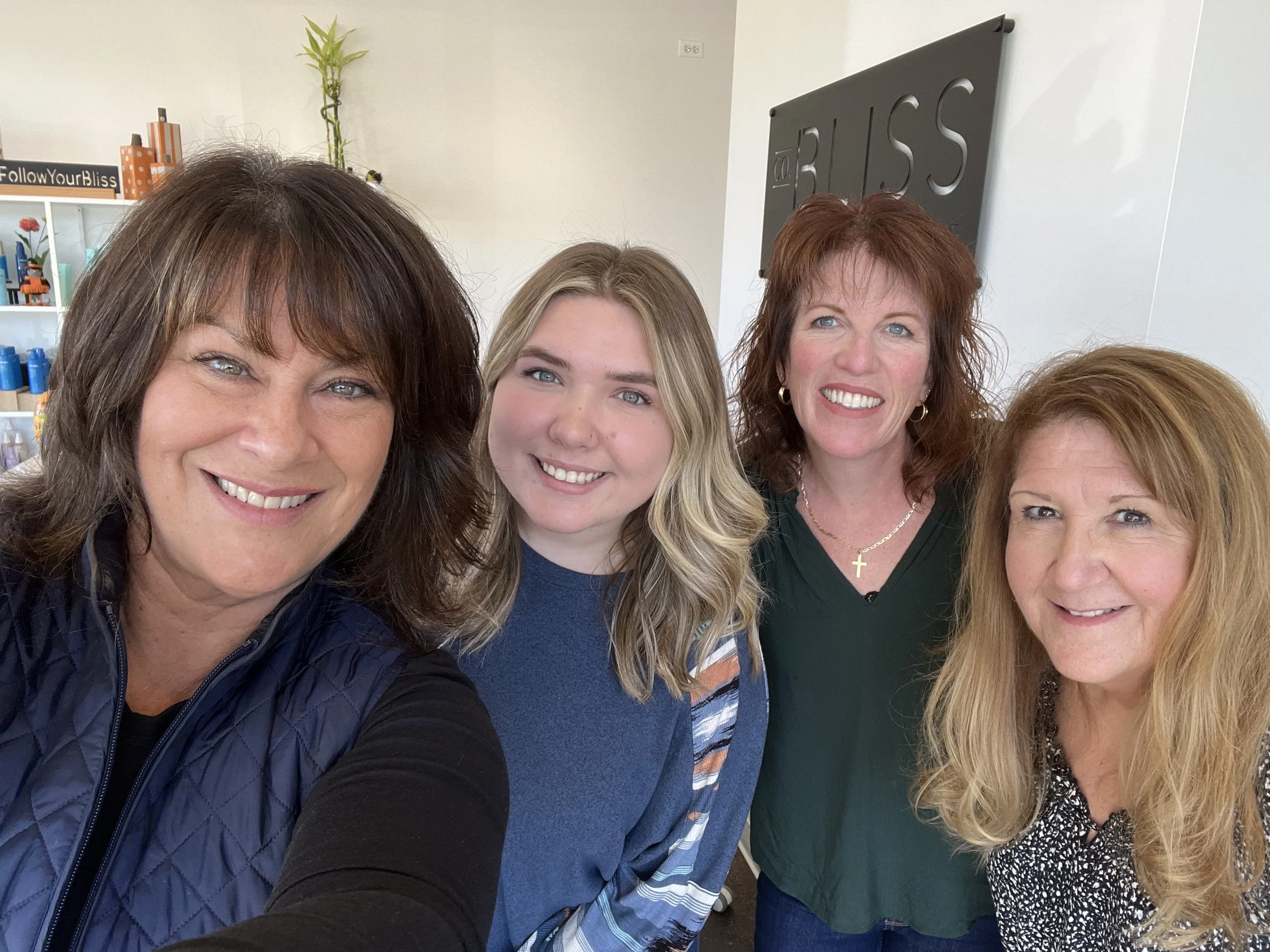 Four women smiling and taking a selfie inside a room with a white wall, shelves with colorful bottles, and a sign that says 'Follow Your Bliss' in the background.