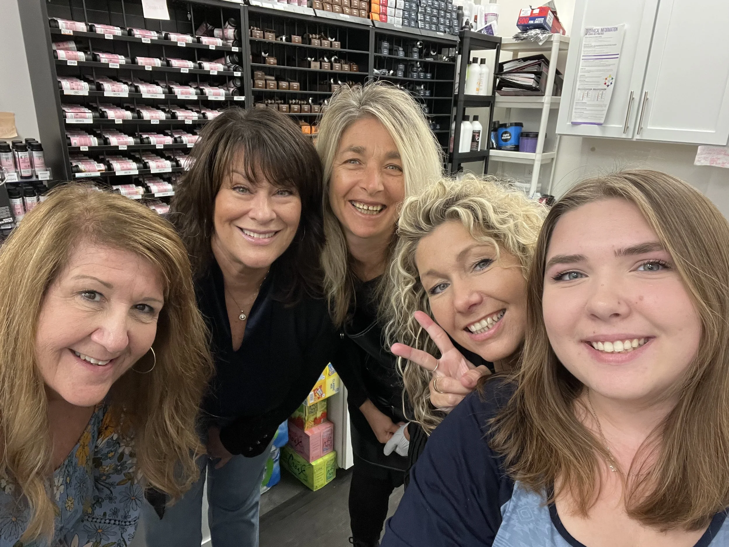 Five women smiling and posing for a selfie inside a salon stockroom with shelves of hair care products in the background.