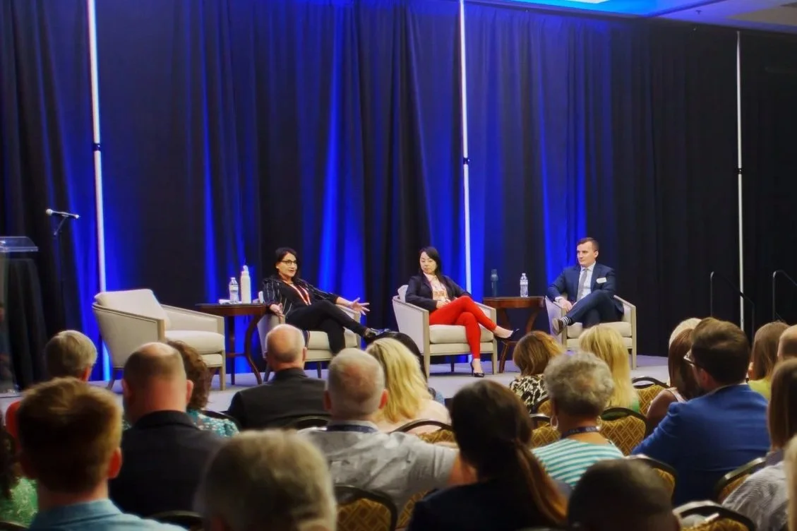 Panel discussion with three people on stage, two women and one man, seated in armchairs on a blue-lit stage with an audience watching.