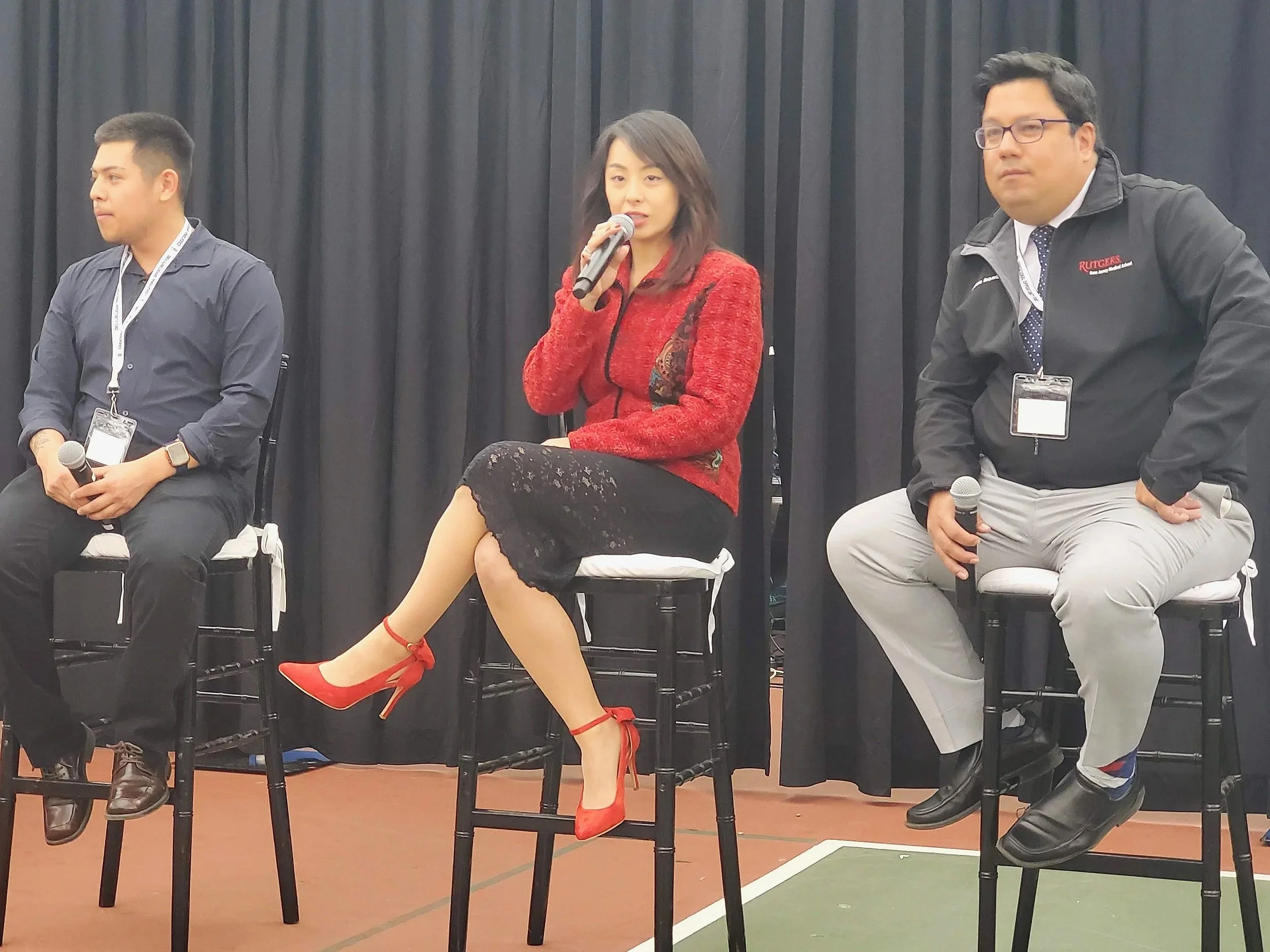 Three people sitting on chairs on a stage with black curtains behind them at a panel discussion, each holding a microphone.