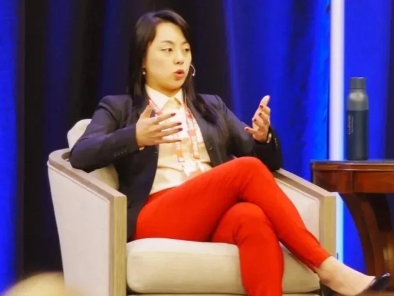 A woman sitting in an armchair, speaking and gesturing with her hands during a panel or interview, with a blue curtain behind her and a modern water bottle on a wooden table beside her.