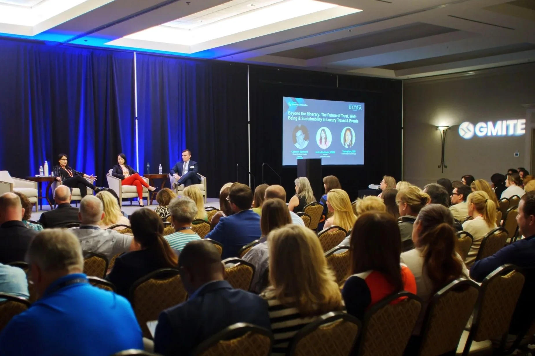 A panel discussion at a conference with three speakers on stage and a large audience seated in front. The stage has a blue backdrop and a screen displaying a presentation titled 'Beyond the Itinerary: The Future of Trust, Well-Being & Sustainability in Luxury Travel & Events.'