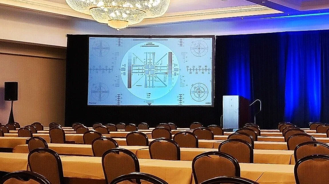 Conference room set up with rows of tables and chairs, a large screen displaying a technical test pattern, and a podium near the stage.