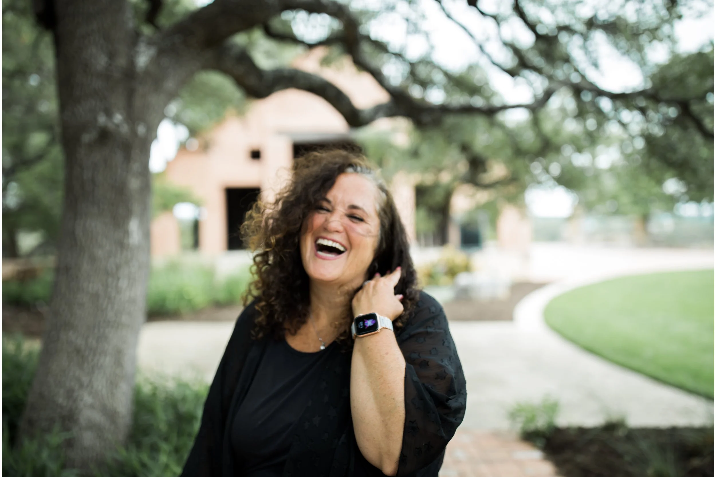 A woman with curly dark hair laughing outdoors, wearing a black top and a smartwatch, standing under a tree with a background of a curved pathway and a building.