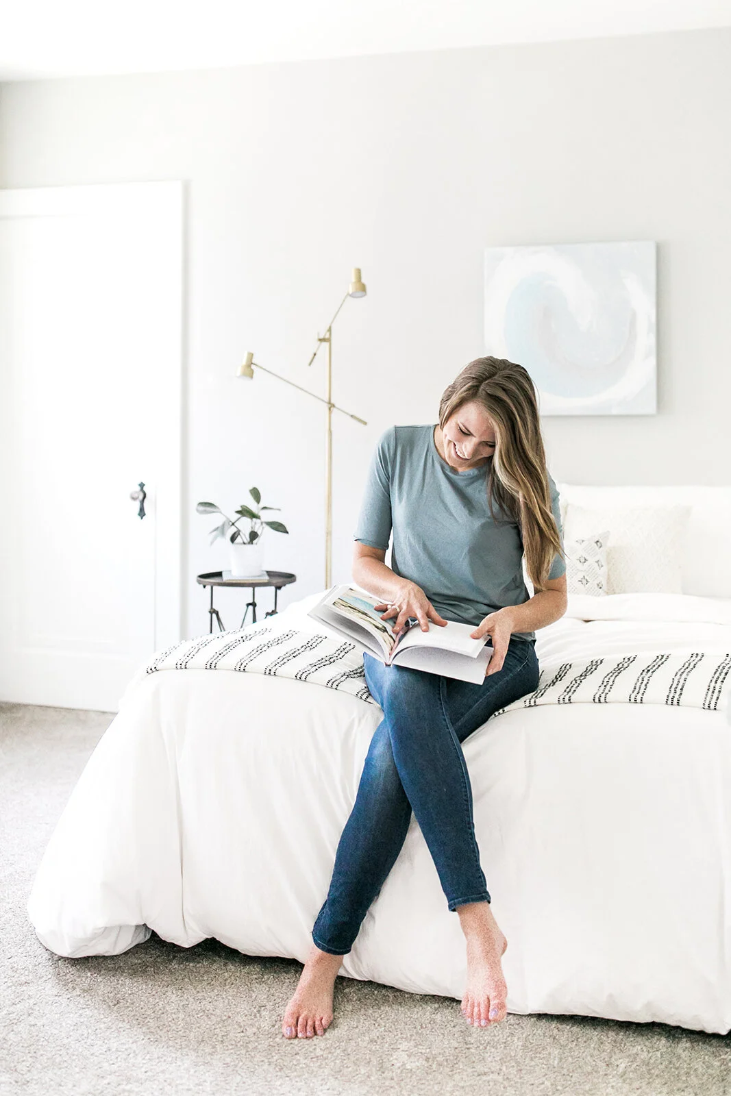A woman with long brown hair sitting on the edge of a white bed, reading a magazine in a bright, minimalist bedroom with white walls and modern decor.