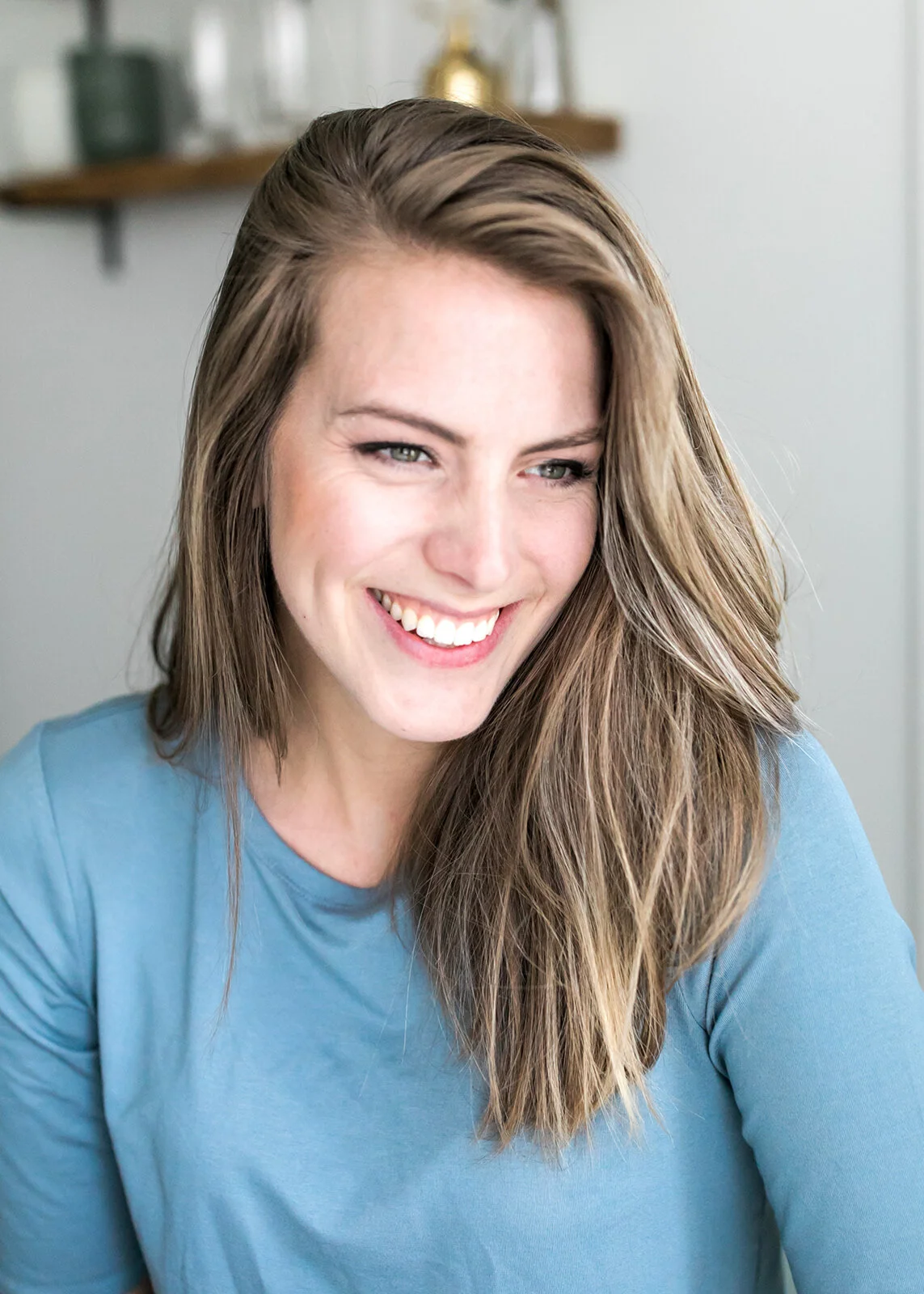 A woman with long, light brown, wavy hair smiling and looking slightly to her right, wearing a blue shirt, in an indoor setting with a blurred background.