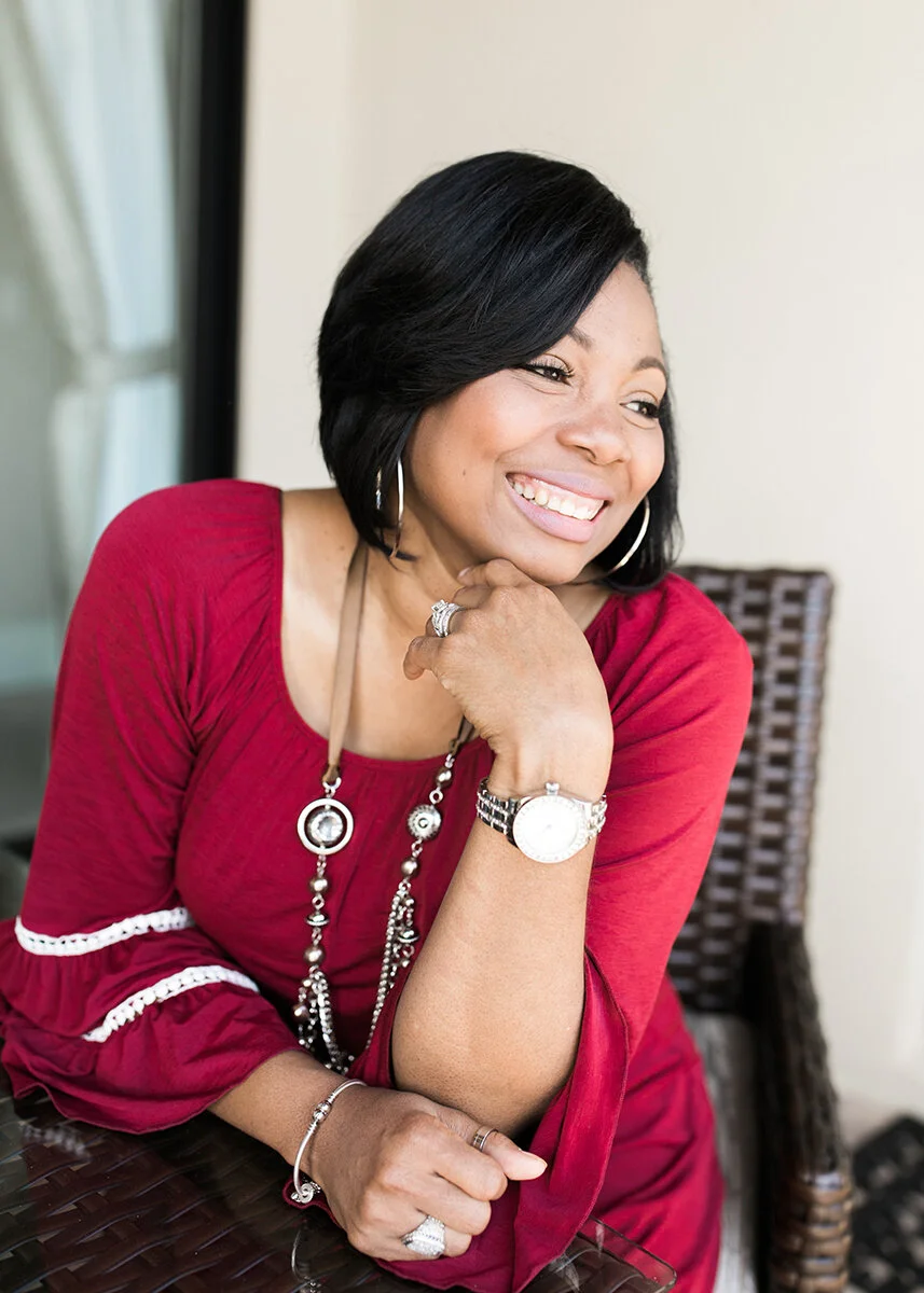 A smiling woman with short black hair, wearing a red dress and jewelry, sitting outdoors and resting her chin on her hand.