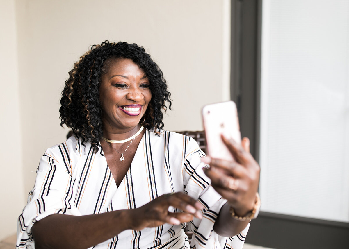 A woman with curly black hair and a striped blouse smiling while taking a selfie with her smartphone in a well-lit room.