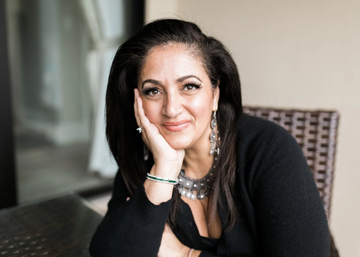 A woman with dark hair, wearing jewelry, resting her head on her hand while sitting at a table indoors.