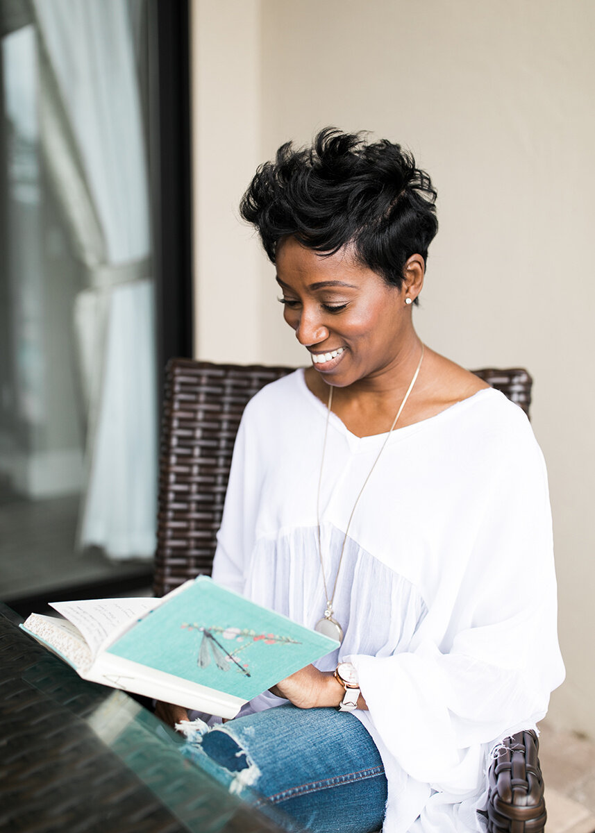 Woman with short curly black hair sitting outdoors, smiling, and reading a book.
