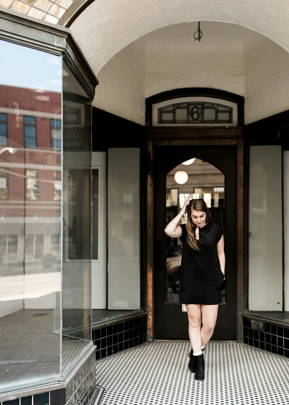 A woman walking through an arched doorway inside a building, wearing a black dress and black ankle boots. Reflections of a brick building are visible in the glass window on the left.