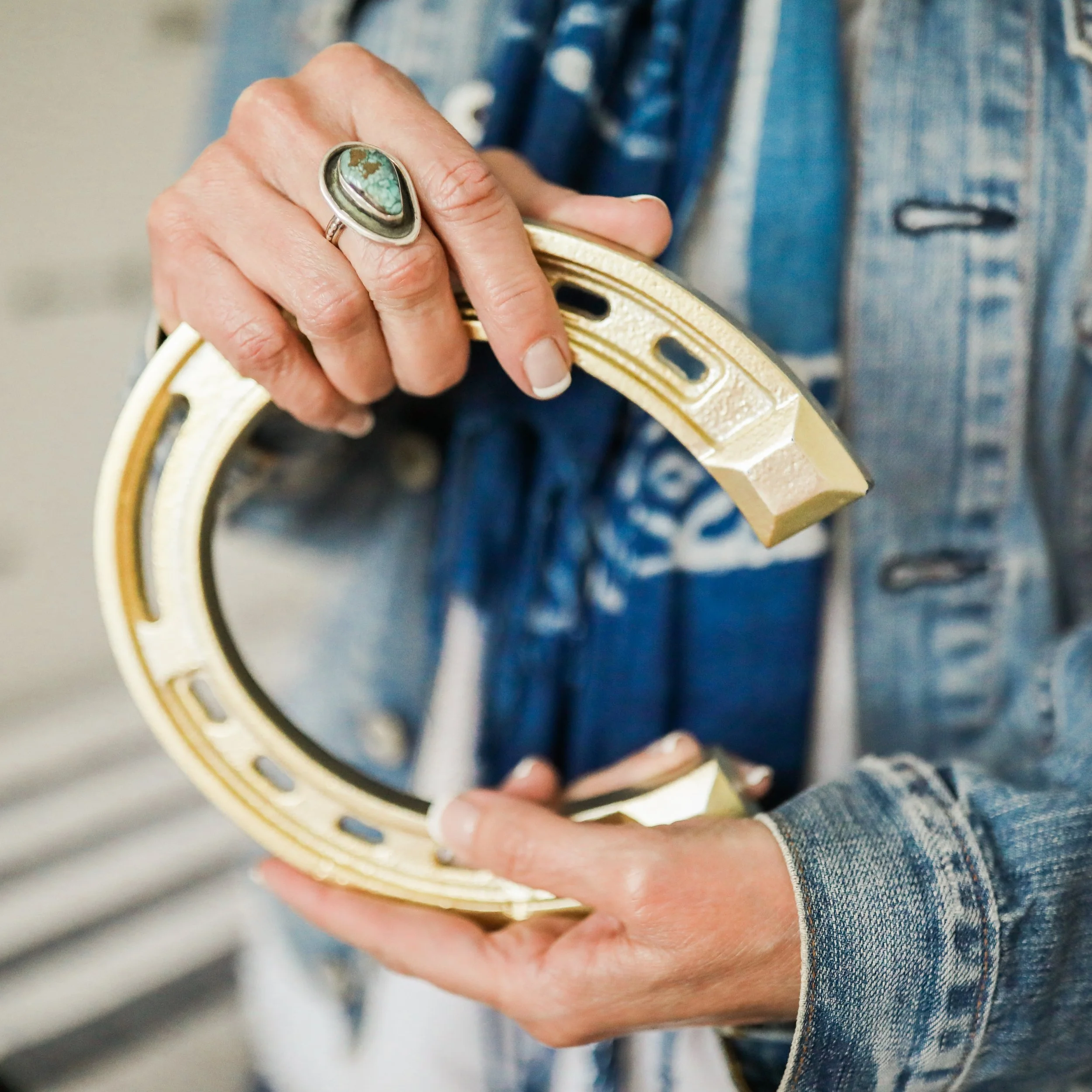 Person holding a shiny, golden horseshoe while wearing a ring with turquoise gemstone and denim jacket.