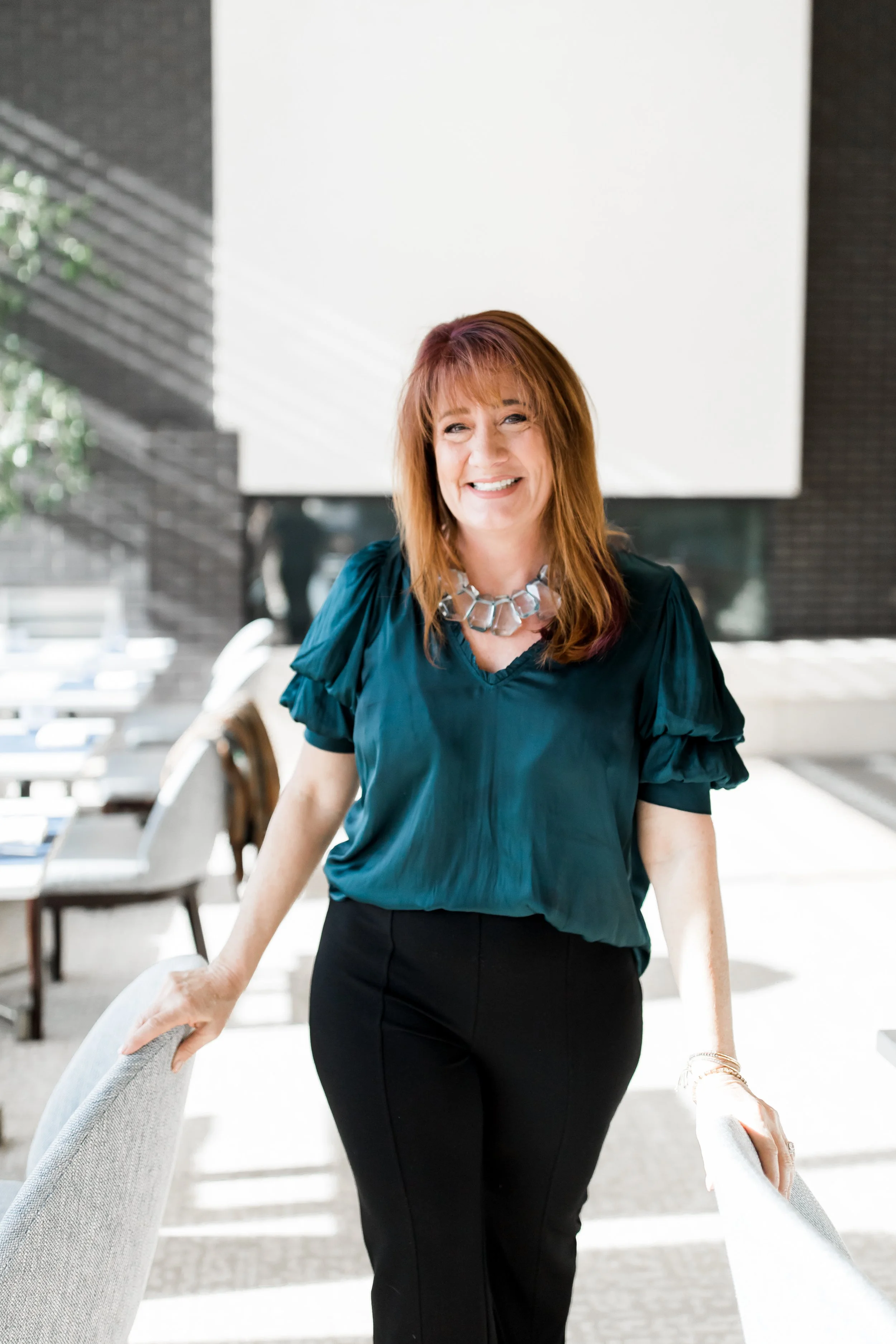 A smiling woman with red hair wearing a teal blouse and black pants, standing in a bright modern indoor space with chairs and tables, holding the back of a chair.