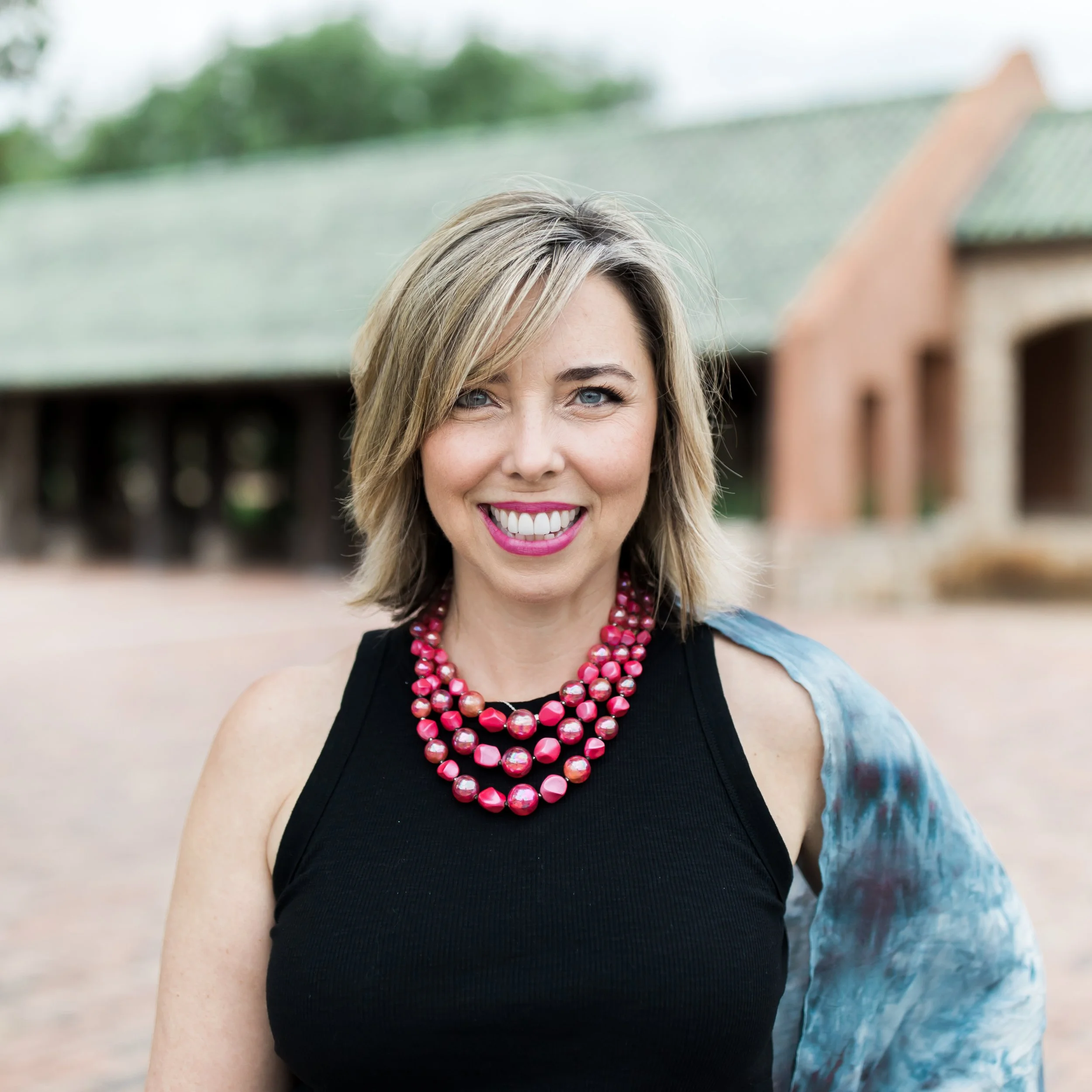 A smiling woman with blonde hair and blue eyes, wearing a black sleeveless top adorned with chunky pink necklaces and a colorful shawl over her shoulder, standing outdoors with a blurred brick building in the background.