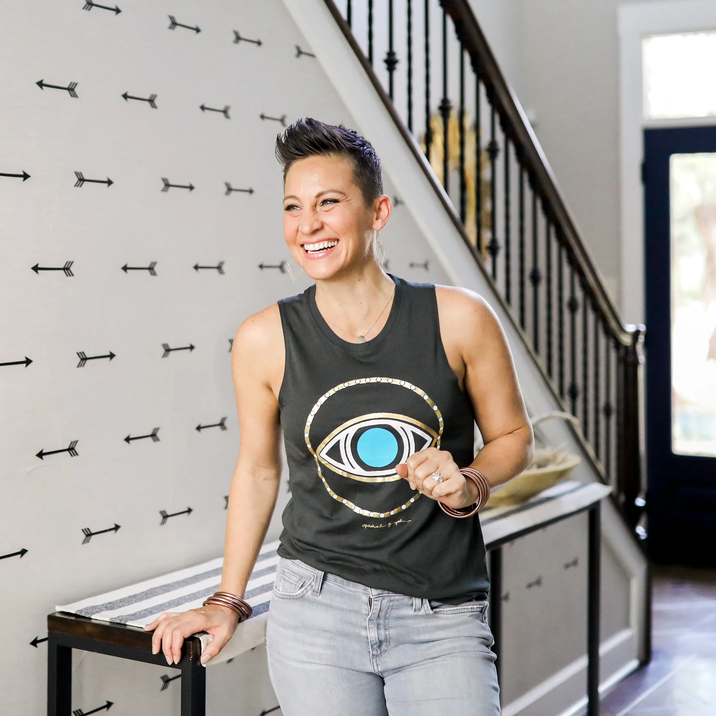 A woman with short dark hair, wearing a black sleeveless top with a large eye design and light gray jeans, smiling and standing indoors near a staircase with black railing and a wall decorated with black arrows.