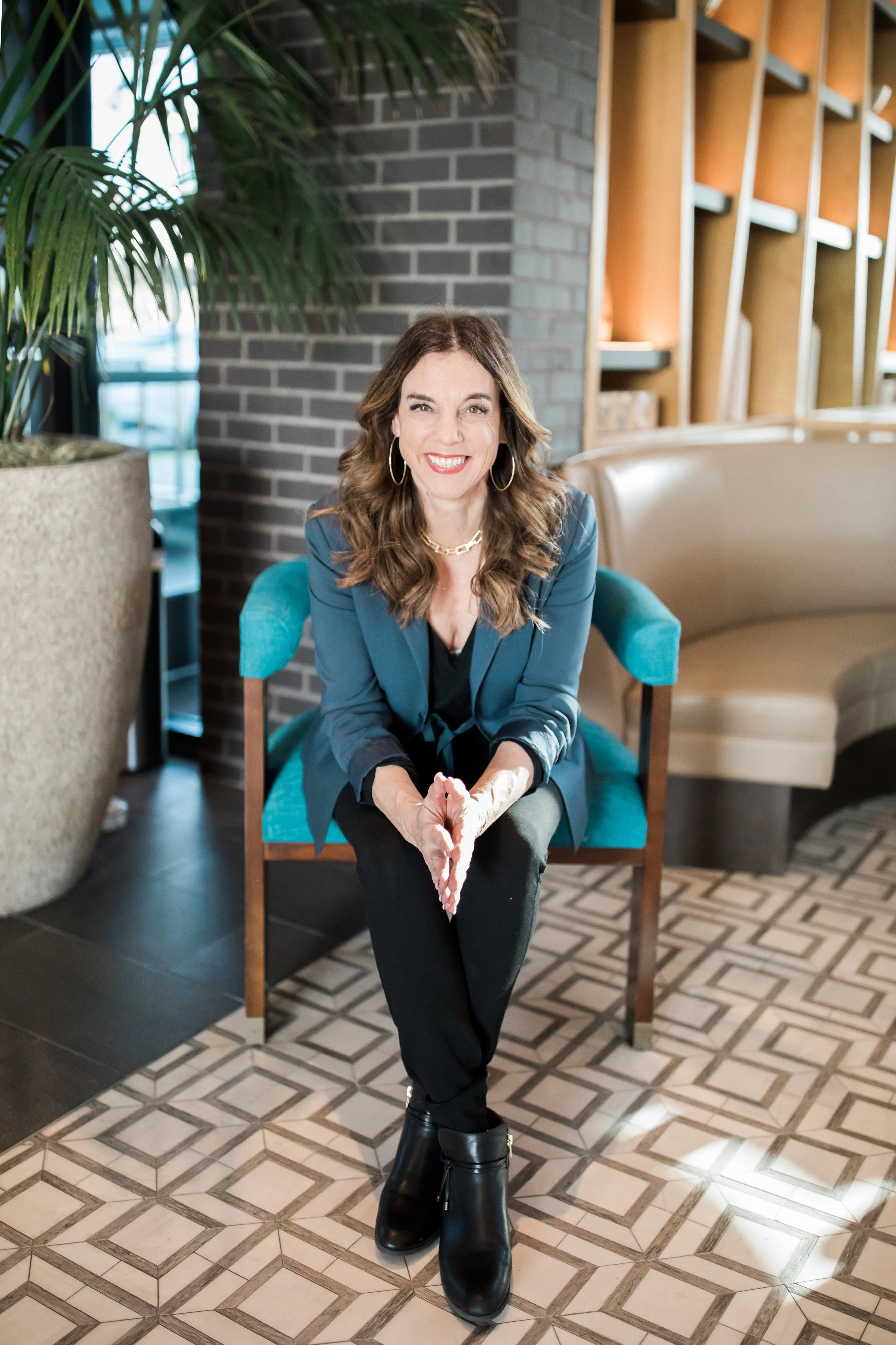 A woman with shoulder-length wavy brown hair, wearing a teal blazer, black top, black pants, and black ankle boots, sits on a wooden chair with teal cushions, smiling at the camera in a modern, stylish interior with a large potted plant, brick wall, 