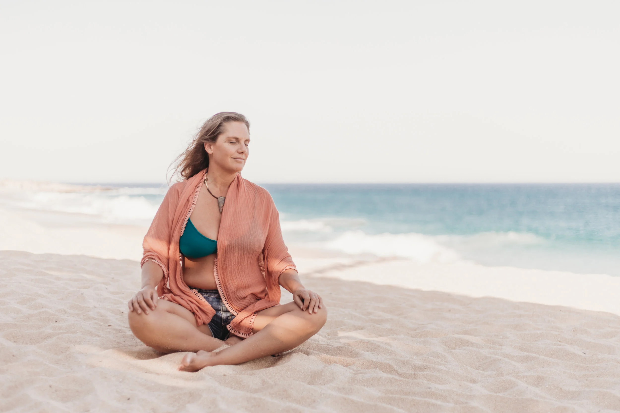 A woman sitting cross-legged on a sandy beach, meditating with her eyes closed, wearing a teal top and an orange open shirt, with the ocean and sky in the background.