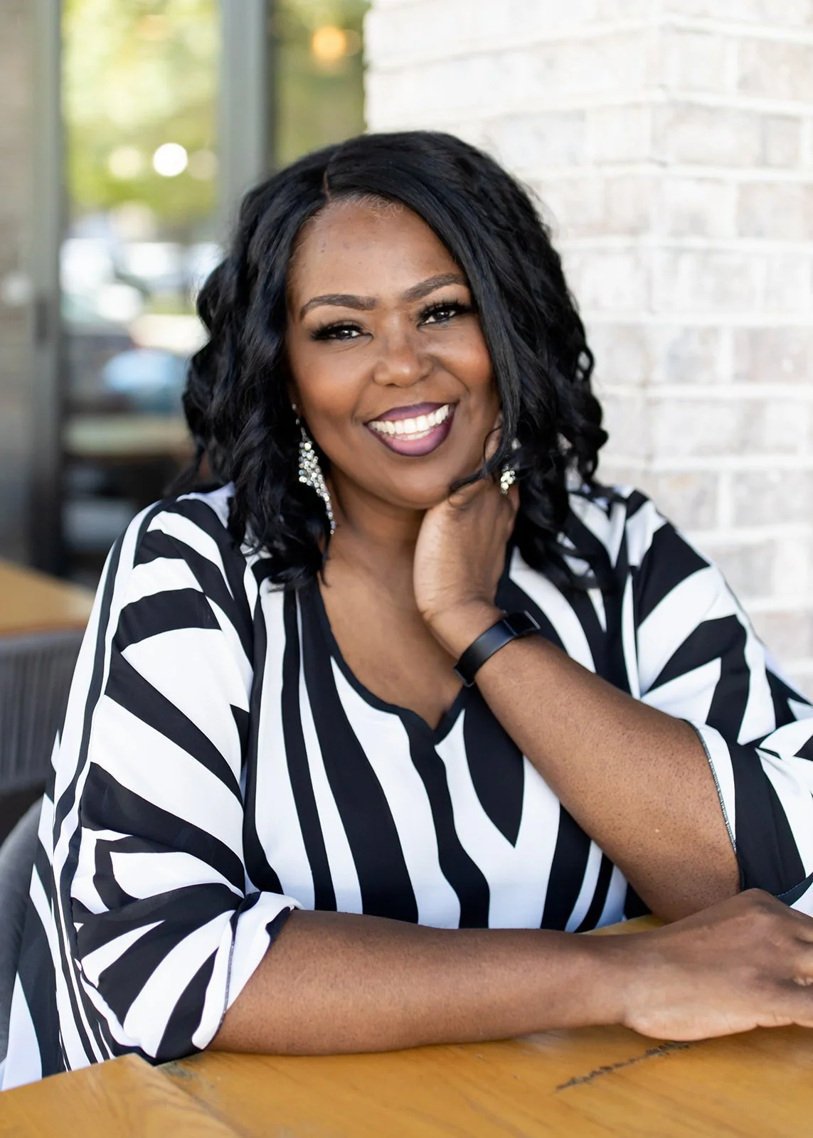 Smiling woman with black curly hair and earrings, wearing a black and white striped top, seated at a wooden table indoors near a brick wall.