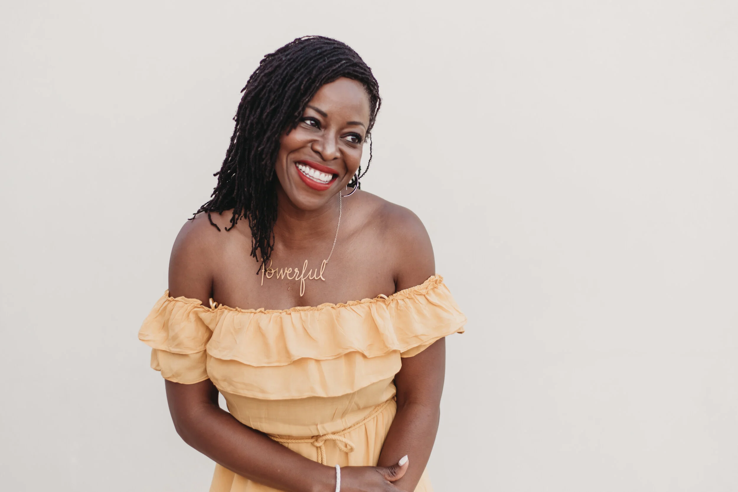 A smiling woman with dark, shoulder-length dreadlocks wearing an off-shoulder yellow dress and a necklace that spells out 'powerful' against a plain white background.