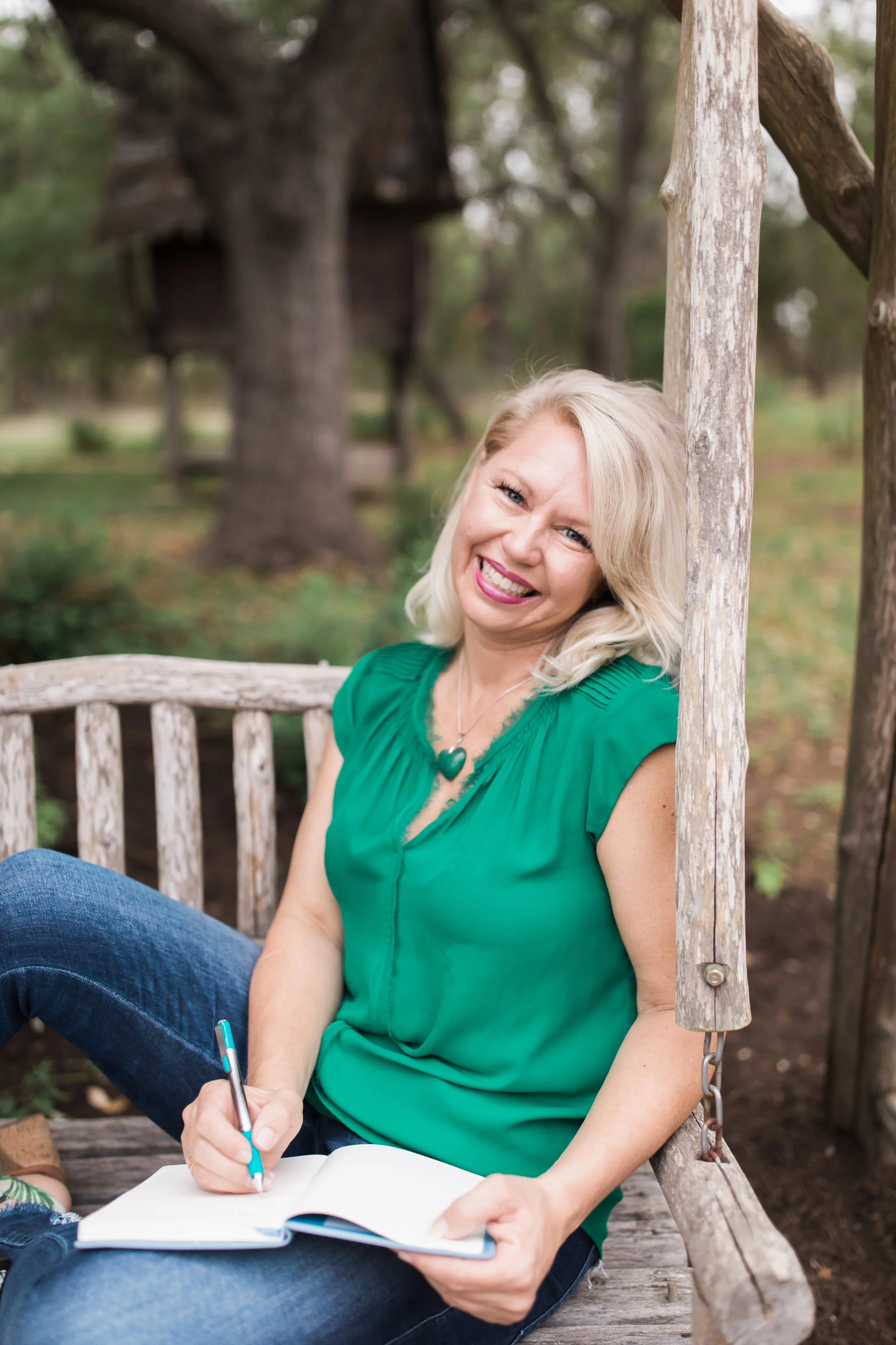 A woman with blonde hair, wearing a green top and jeans, sitting on a wooden swing outside, smiling and holding a pen and a notebook.